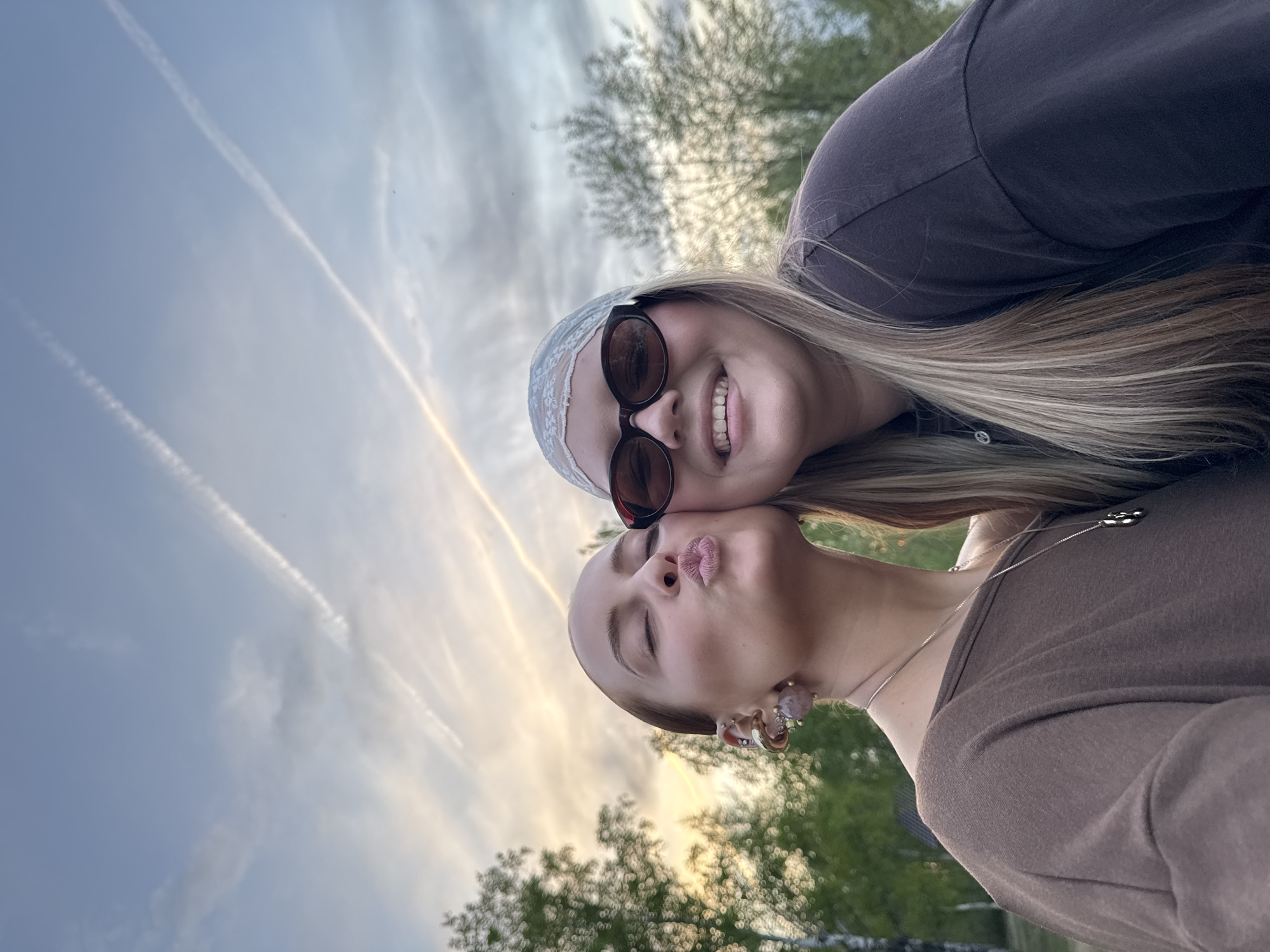Two Caucasian women pose for a selfie outdoors. One woman is kissing the other on the cheek. The woman being kissed wears sunglasses and a headscarf. The background includes trees and a cloudy sky with contrails. The exact location is unclear, and cannot be determined as Chaikovsky, Russia.