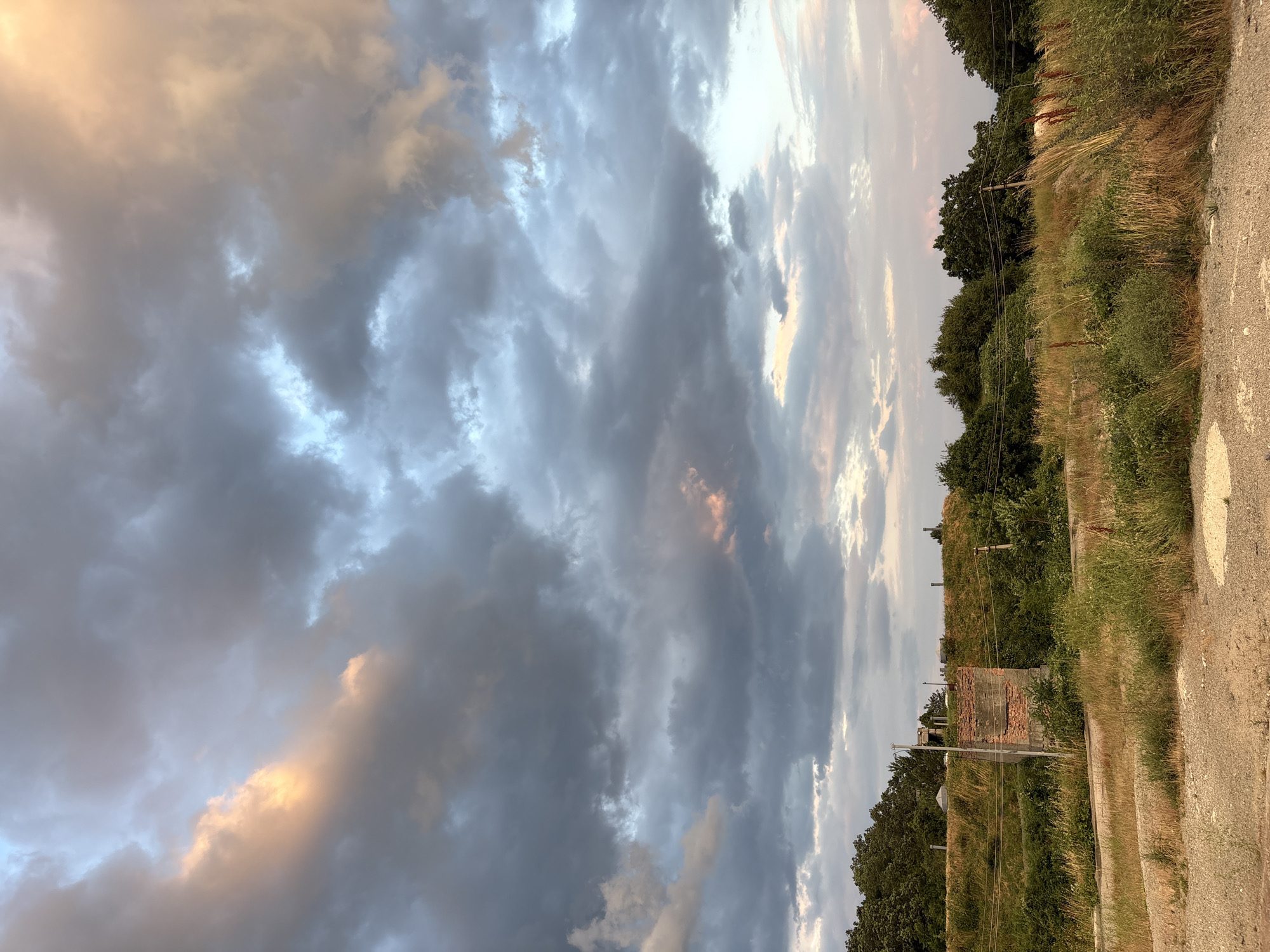 This is a photograph of a landscape in Malyi Mayak, Ukraine, likely taken at dusk. The sky is filled with dramatic, textured clouds, with patches of soft orange and pink light breaking through the gray and blue tones. The clouds suggest either the end of a stormy day or the approach of one. Below the sky, a line of trees forms the horizon, with lush green foliage on the left transitioning to a more sparse, wilder vegetated area in the center and right. In the foreground, there is dry, brown grass and dirt. A partially constructed brick structure, possibly a remnant of a building, stands prominently to the left of center, adjacent to a patch of overgrown vegetation. Power lines are visible stretching across the scene, supported by poles. The overall impression is one of a rural or semi-abandoned area under a dynamic, colorful sky.