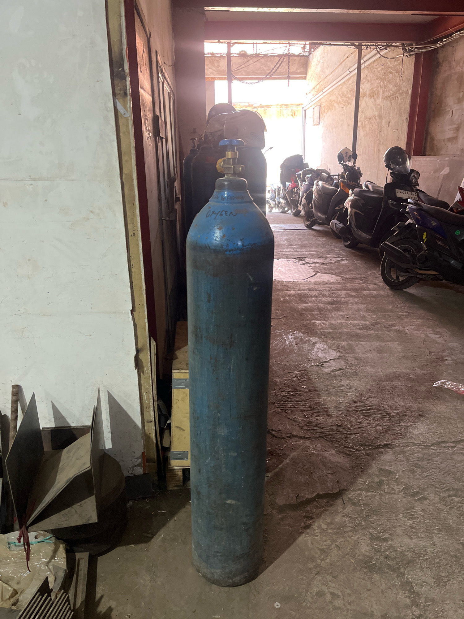 This is a forensic, matter-of-fact description of a scene in a garage or workshop, likely located in Cimahi, Indonesia, given the presence of numerous motorcycles.
The primary subject is a large, blue oxygen tank positioned prominently in the foreground, slightly to the right of center. The tank is cylindrical and appears to be made of metal, with a valve mechanism visible at the top. Faint lettering, appearing to spell "OXYGEN," is visible on the side of the tank. The tank casts a distinct shadow on the concrete floor.
To the left of the oxygen tank, a portion of a white, textured wall is visible, with a vertical, dark-colored edge running alongside it. Further to the left and partially obscured by the oxygen tank, several metallic objects are arranged. These appear to be flat, angular pieces of metal, some of which are bound together with red cord. Beneath these, other items are wrapped in plastic sheeting.
Behind the oxygen tank, the scene opens up into a wider space that appears to be an alleyway or covered walkway. A row of parked motorcycles is lined up along the right side of this space. The motorcycles are of various colors and models, and several have helmets placed on them. The overall environment is dimly lit, with sunlight streaming in from an opening at the far end of the space, creating bright highlights and shadows on the concrete floor and walls. The walls themselves appear to be constructed of concrete or stucco and show signs of age and wear. Structural elements, possibly beams, are visible overhead. The ground is a rough concrete surface with visible cracks and discoloration.