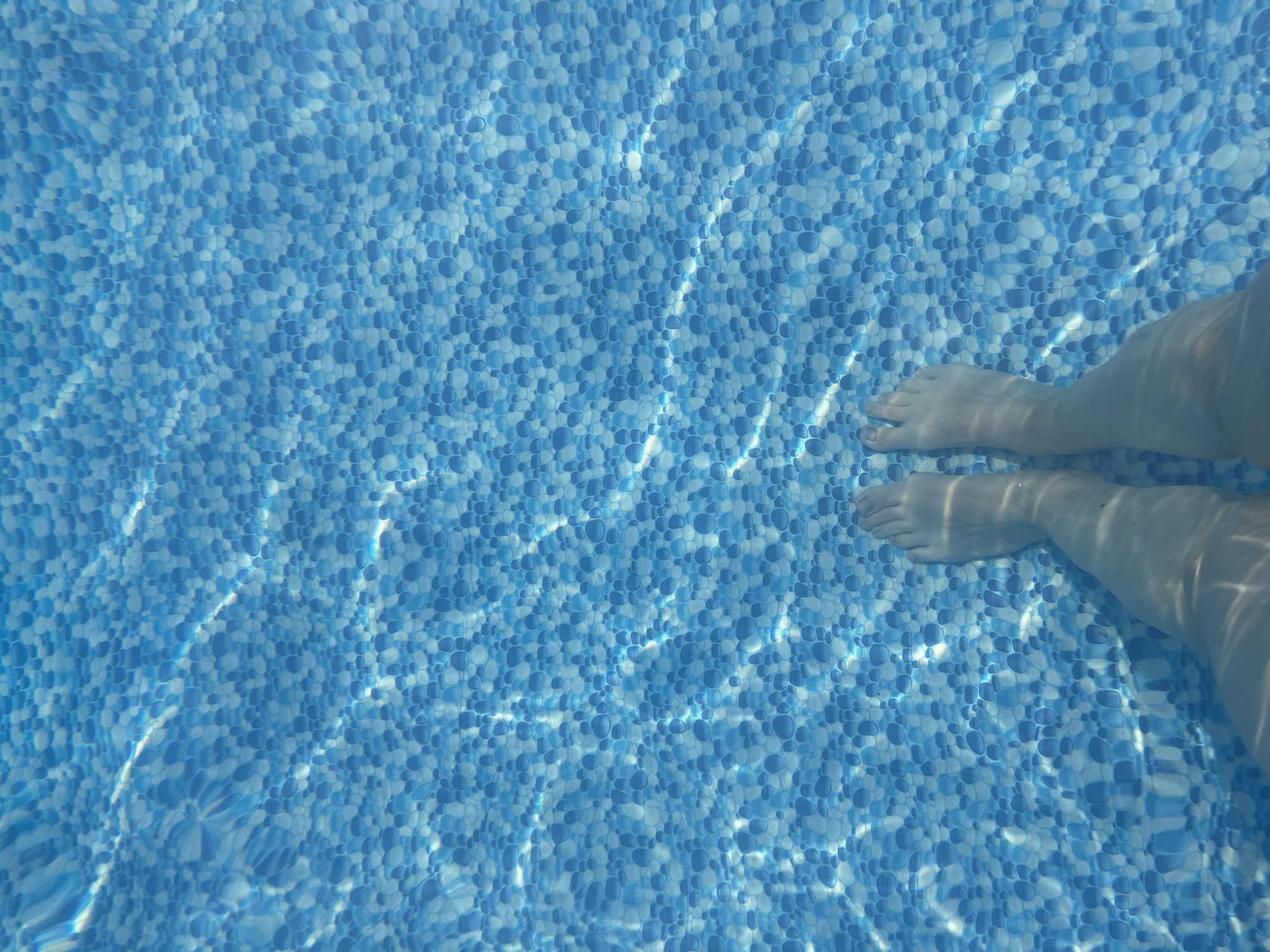 The image shows a person's feet submerged in a swimming pool. The pool has a blue, pebble-like pattern on the bottom. Sunlight creates shimmering patterns on the water's surface, refracting through the water and casting light and shadow on the pool floor. The water appears clear, allowing the pattern on the bottom to be clearly visible. The perspective is from above, looking down at the feet. There is no information in the image to suggest a specific location within the city of Orenburg, Russia.