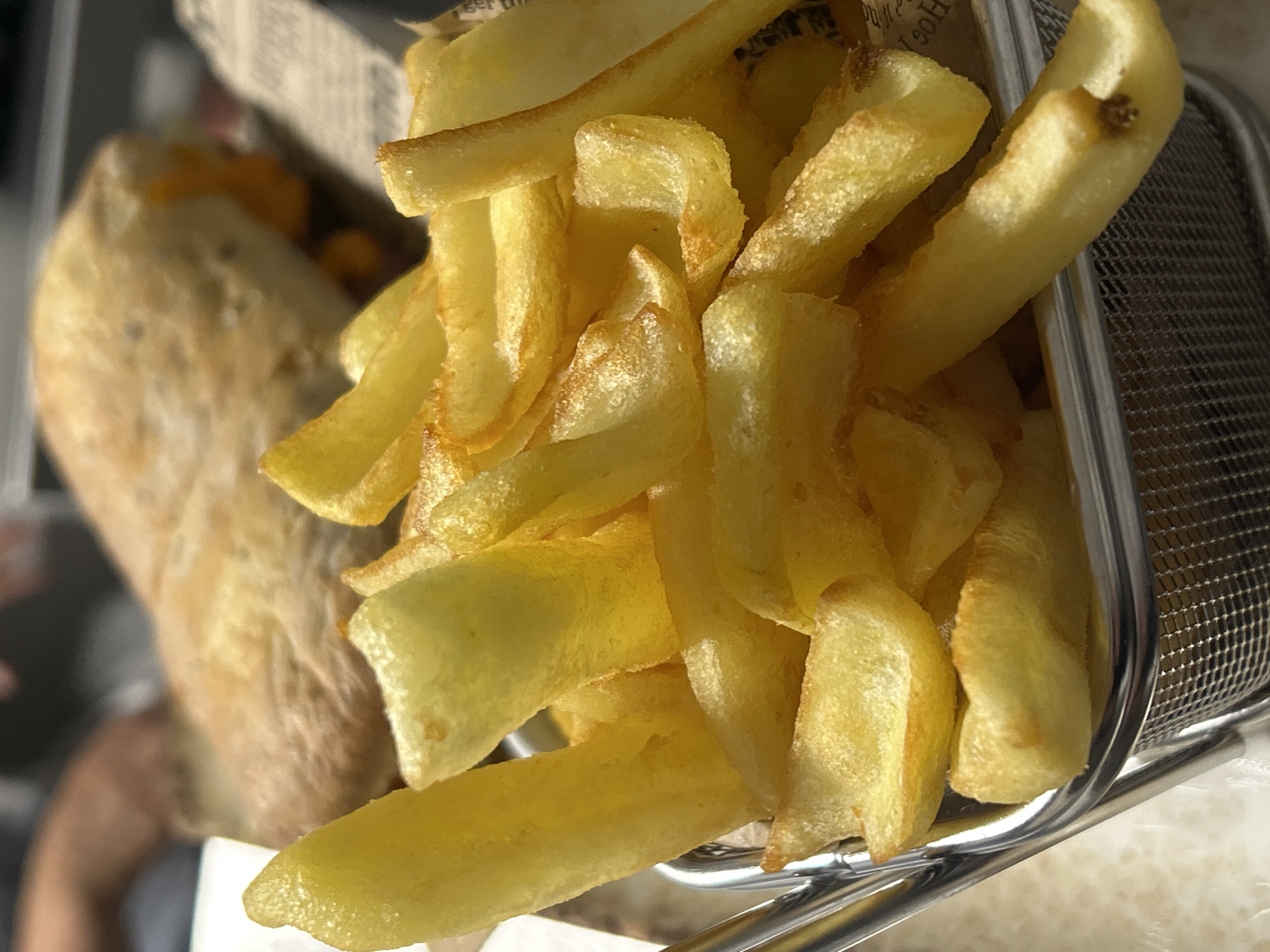 A close-up shot features a metal basket overflowing with golden-yellow french fries. To the left, partially out of focus, is a sandwich in packaging. The fries appear to be freshly cooked with a slightly uneven coloration. The scene likely takes place in a restaurant or cafe setting, although the location in Burgas, Bulgaria, is not determinable from the image alone. The only objects are the fries, basket, and the sandwich. There are no people visible in focus, only the partially-visible arm of a person out of focus in the bottom left corner. There are no visible actions or interactions. The environment is neutral and undefined.