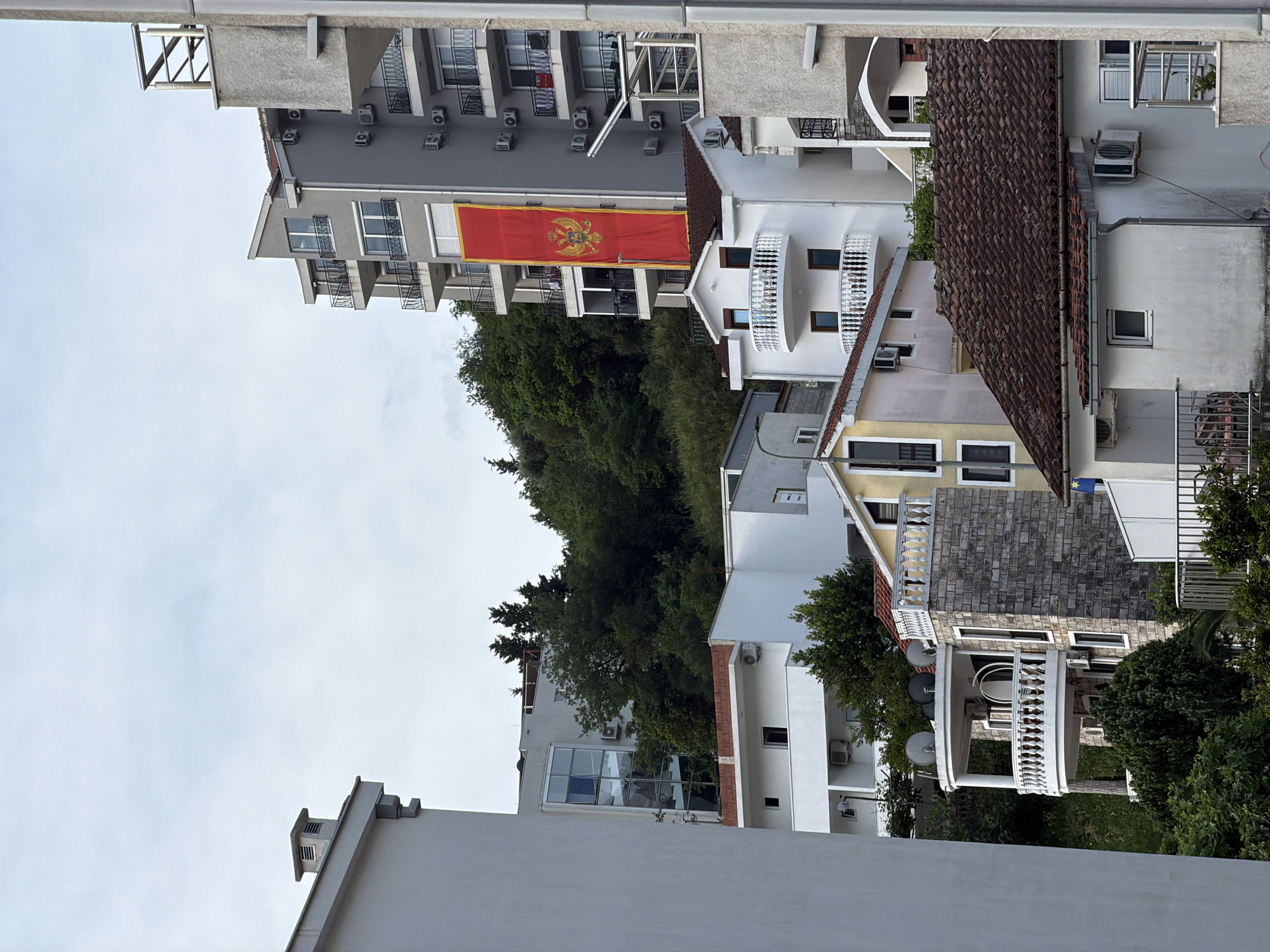 The image shows a view of buildings and trees in Budva, Montenegro. The perspective is slightly elevated, looking down on the scene. The sky is overcast. Several buildings of different heights and architectural styles are visible. One building has a large Montenegrin flag hanging from it. Other details include balconies, air conditioning units, and satellite dishes on some of the buildings. Trees are interspersed between the buildings, adding greenery to the scene. The location appears to be a residential area within Budva.