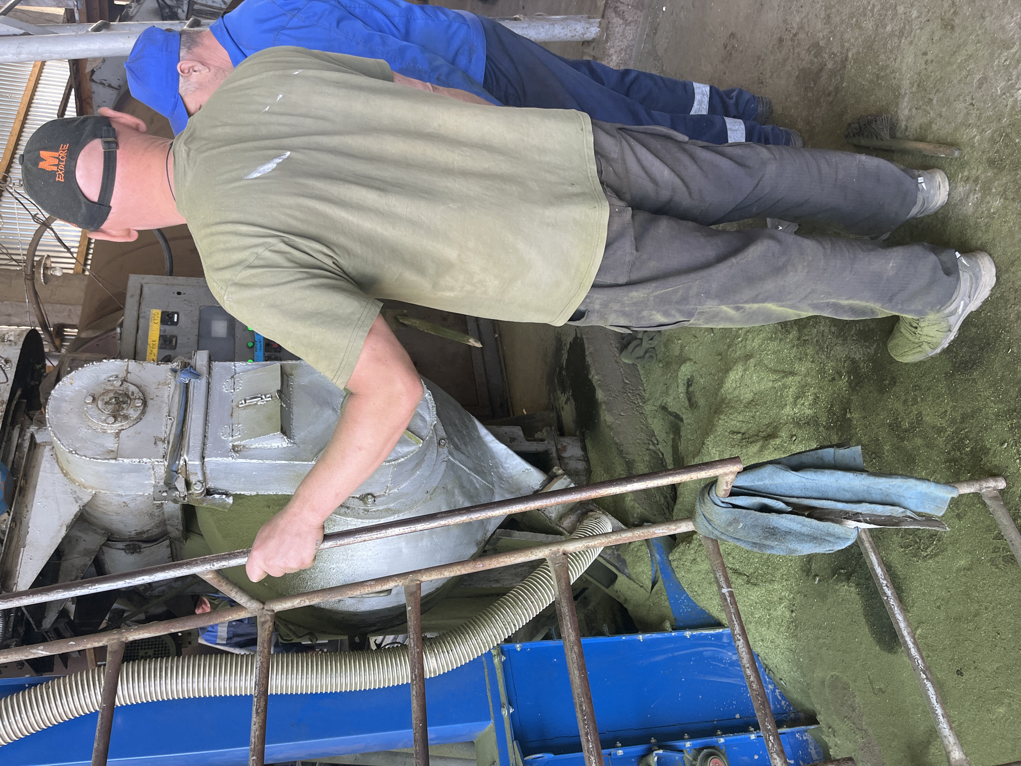 The photograph depicts two individuals in an industrial environment, likely indoors, as suggested by the presence of heavy machinery and metal structures. One person is wearing a black baseball cap with an orange "M" logo and the word "EXPLORE" on it, a green t-shirt, and gray work pants, while the other person is wearing a blue shirt and a blue cap. They are standing next to a large, silver-colored piece of machinery, possibly agricultural equipment. The area surrounding the machine is covered in a green particulate matter. A blue rag is draped over a metal railing. The location and city are not discernible from the visual information provided in the image.