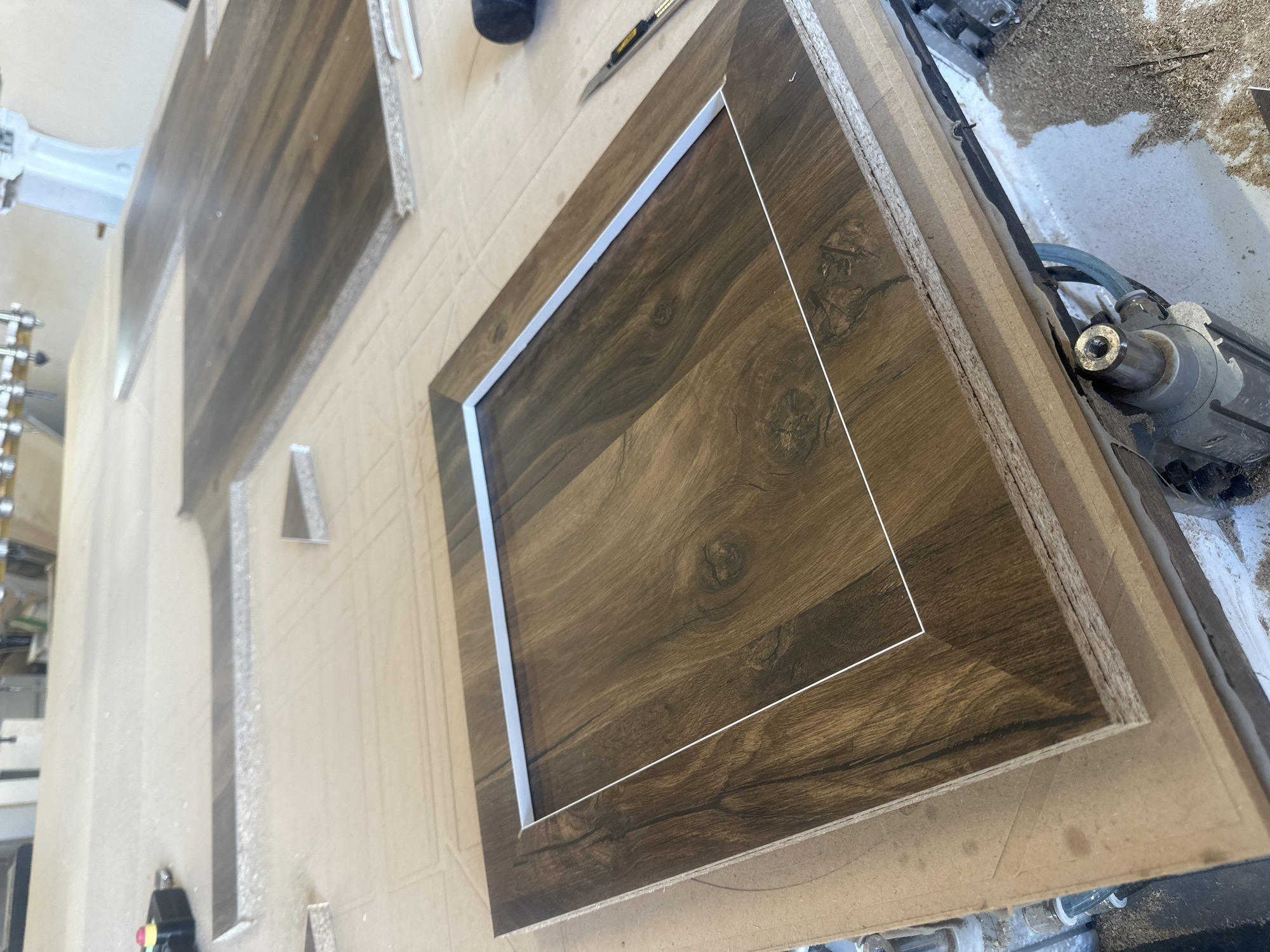 The image shows a close-up of woodworking on a CNC machine. Visible are pieces of wood with a brown wood-grain laminate. One piece has a square opening with a white liner. The machine itself and its components are partially visible, as is some wood debris. The location is not discernible from the image. There are no people visible. There is no indication that this image was taken in Kalimantsi, Bulgaria.