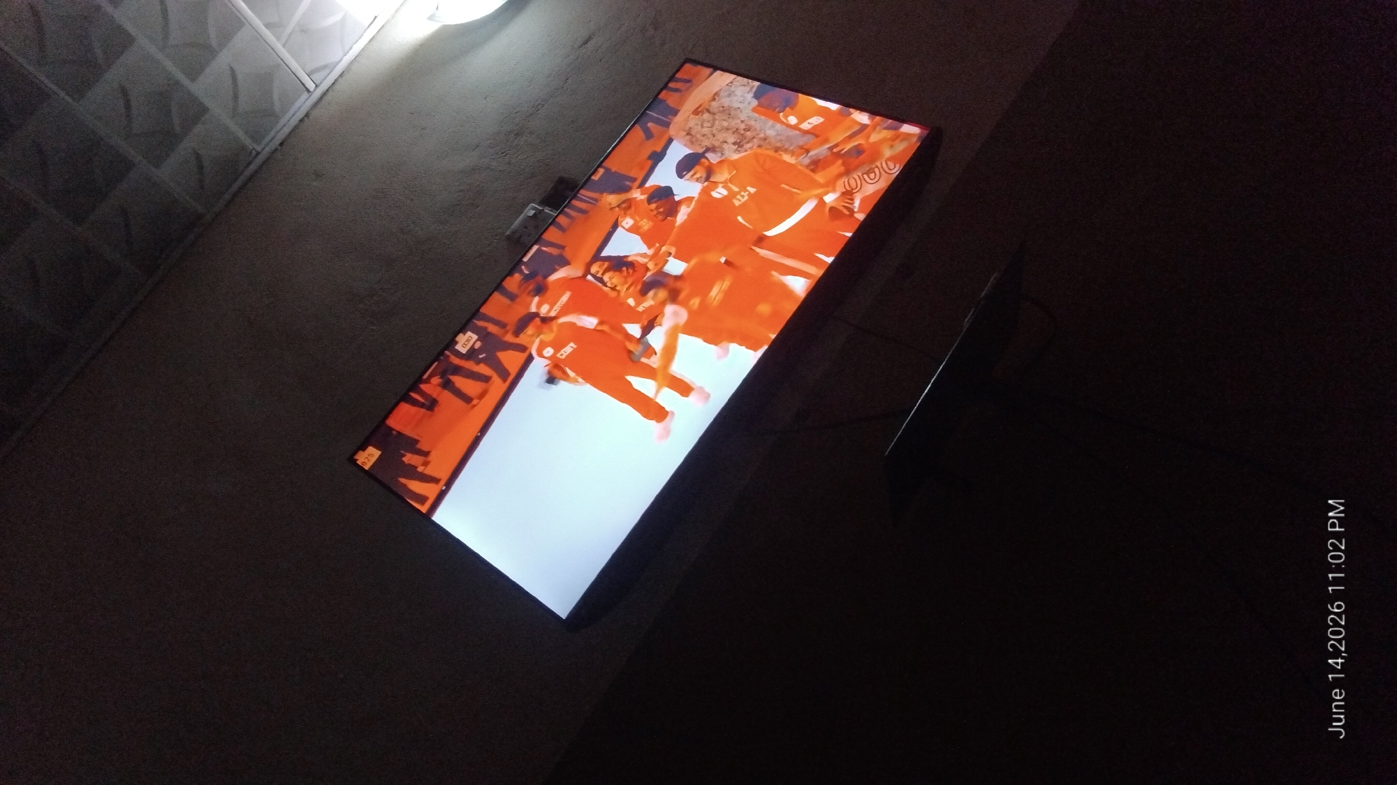 The image shows a television screen displaying a group of people in orange uniforms, possibly a sports team or performers. The room is dimly lit, with the ceiling tiles visible and appearing to be made of a standard suspended ceiling material. The television is mounted on a wall. The timestamp in the bottom left corner indicates "June 14, 2026 11:02 PM". The image does not provide any information about the specific location within the city of Howayi, Nigeria.