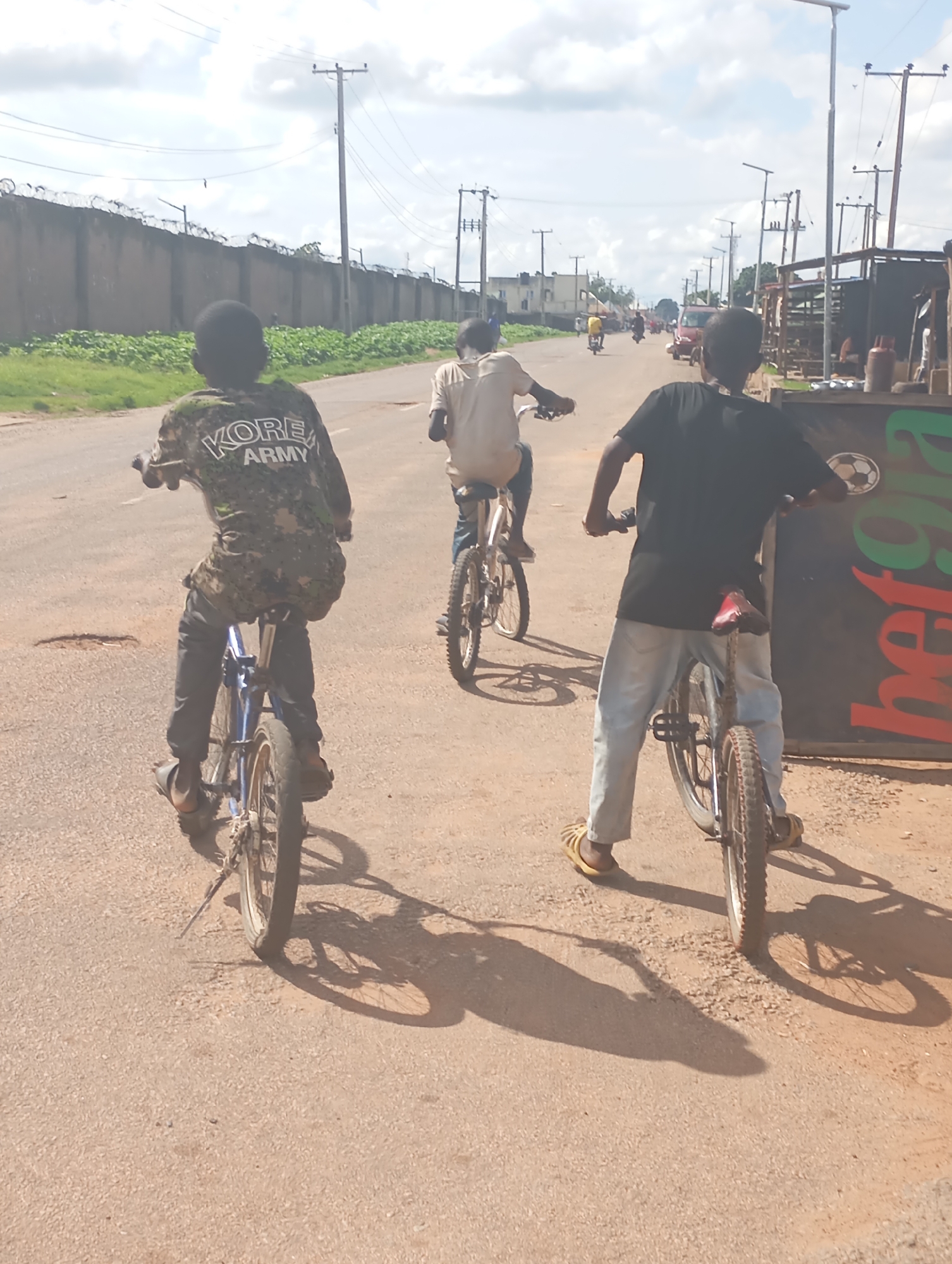 Three young people are riding bicycles down a dusty, paved road in Shendam, Nigeria. The sun is high in the sky, casting long shadows. The weather appears to be clear and warm.
The person in the foreground on the right is wearing a black t-shirt and light blue jeans, with yellow sandals. They are riding a bicycle with a red seat.
The person in the middle is wearing a light-colored t-shirt and dark pants. They are riding a bicycle and appear to be looking ahead.
The person on the left is wearing a camouflage-patterned shirt with "KORE ARMY" printed on the back in white letters, and dark pants. They are riding a blue bicycle.
The road is lined with buildings and vegetation. On the left, there is a long, high wall topped with barbed wire, behind which green bushes are visible. On the right, there is a stall or shop with a dark sign that has green and red lettering. The lettering appears to include the word "herg" or "herg-a". There are also power lines and utility poles visible throughout the scene. Other vehicles and people can be seen in the distance, suggesting it is a busy area. The overall mood appears to be casual and leisurely, with the subjects enjoying a bike ride.