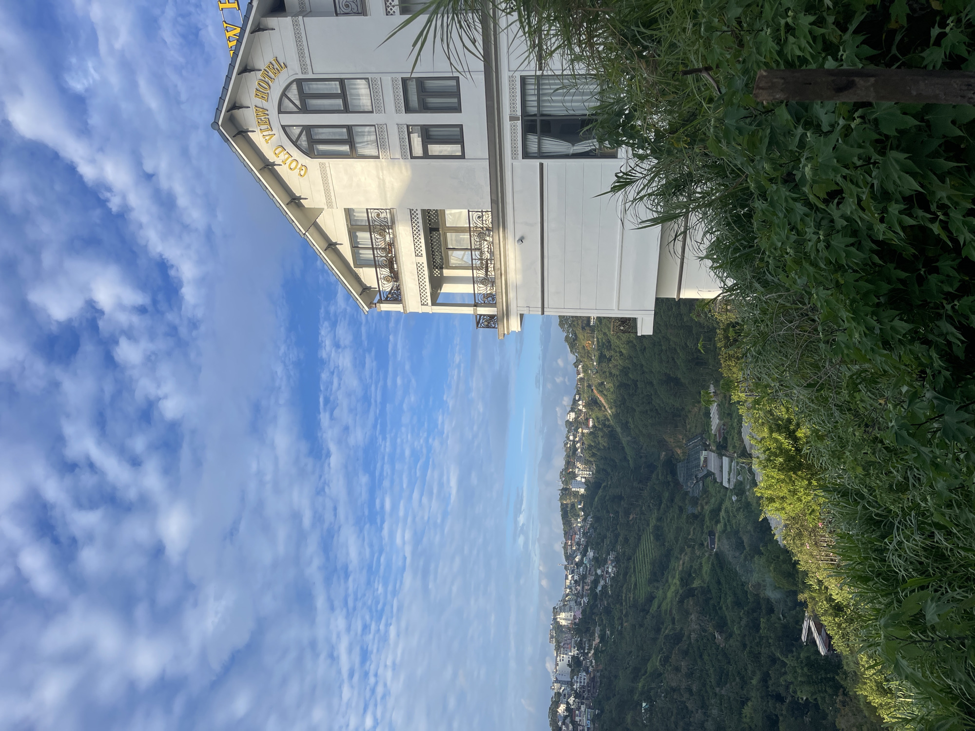 The image is a scenic view of the Gold View Hotel in Da Lat, Vietnam, taken from a slightly elevated perspective. The hotel, a white multi-story building with a peaked roof, is prominently featured on the right side of the frame. It has several balconies with ornate railings and large windows. The words "GOLD VIEW HOTEL" are visible in gold lettering on the hotel's facade.
Below and to the left of the hotel, a sprawling city of Da Lat unfolds, characterized by numerous buildings of varying sizes and architectural styles, nestled into the lush, green hills. The landscape is a mix of dense forestation, terraced farms, and some greenhouses in the distance. The sky above is a vibrant blue, filled with scattered, white, wispy clouds. In the foreground, tall green foliage and grass partially obscure the view, with a wooden post visible on the right. The lighting suggests it is daytime, possibly morning or late afternoon, given the soft sunlight illuminating the scene.