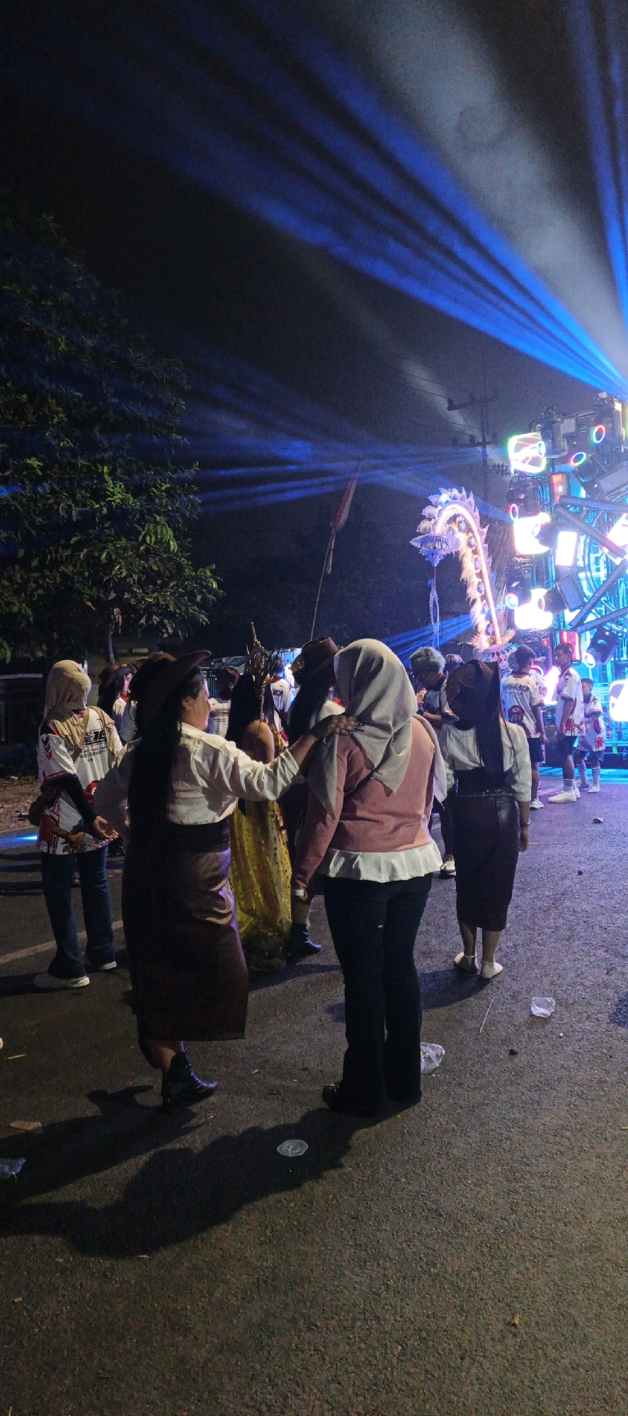 In Kasiyan, Indonesia, a nighttime outdoor event is in progress, illuminated by bright blue stage lights that streak across a dark sky. The scene focuses on a group of people gathered on a paved surface, likely a street or open area. Several individuals are wearing traditional Indonesian attire, including women in headscarves and long skirts. One woman, in the foreground and facing away from the camera, wears a brown cowboy hat, a white shirt, and a brown skirt, with her left hand resting on the shoulder of another woman. This second woman, also facing away, is wearing a light pink top and dark pants, with a grey headscarf. Behind them, a large, elaborate dragon puppet, likely part of a parade or performance, is suspended and illuminated. Further back, a stage setup with multiple lights and speakers is visible. The ground is littered with some debris, suggesting it is a public space. The overall atmosphere suggests a festive cultural celebration.