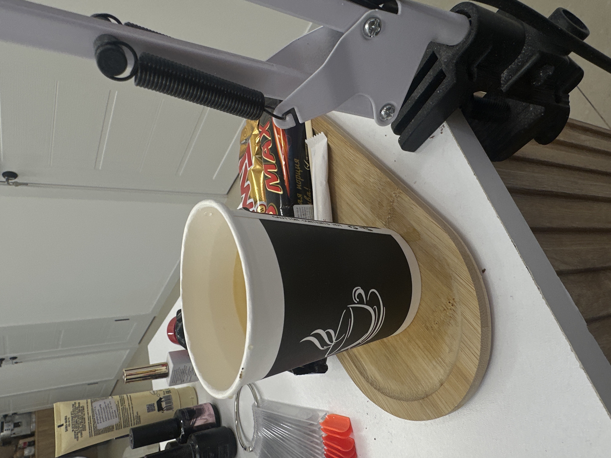 The image shows a white desk with a cup of coffee resting on a wooden tray. Behind the cup are candy bars and a white packet. To the left of the cup are various cosmetics, including nail polish and lotion. Above the scene is a white desk lamp with a black mounting fixture. The background includes white cabinetry. There are no people visible. The location is not specified, and the city of Yerevan, Armenia cannot be determined from the image.