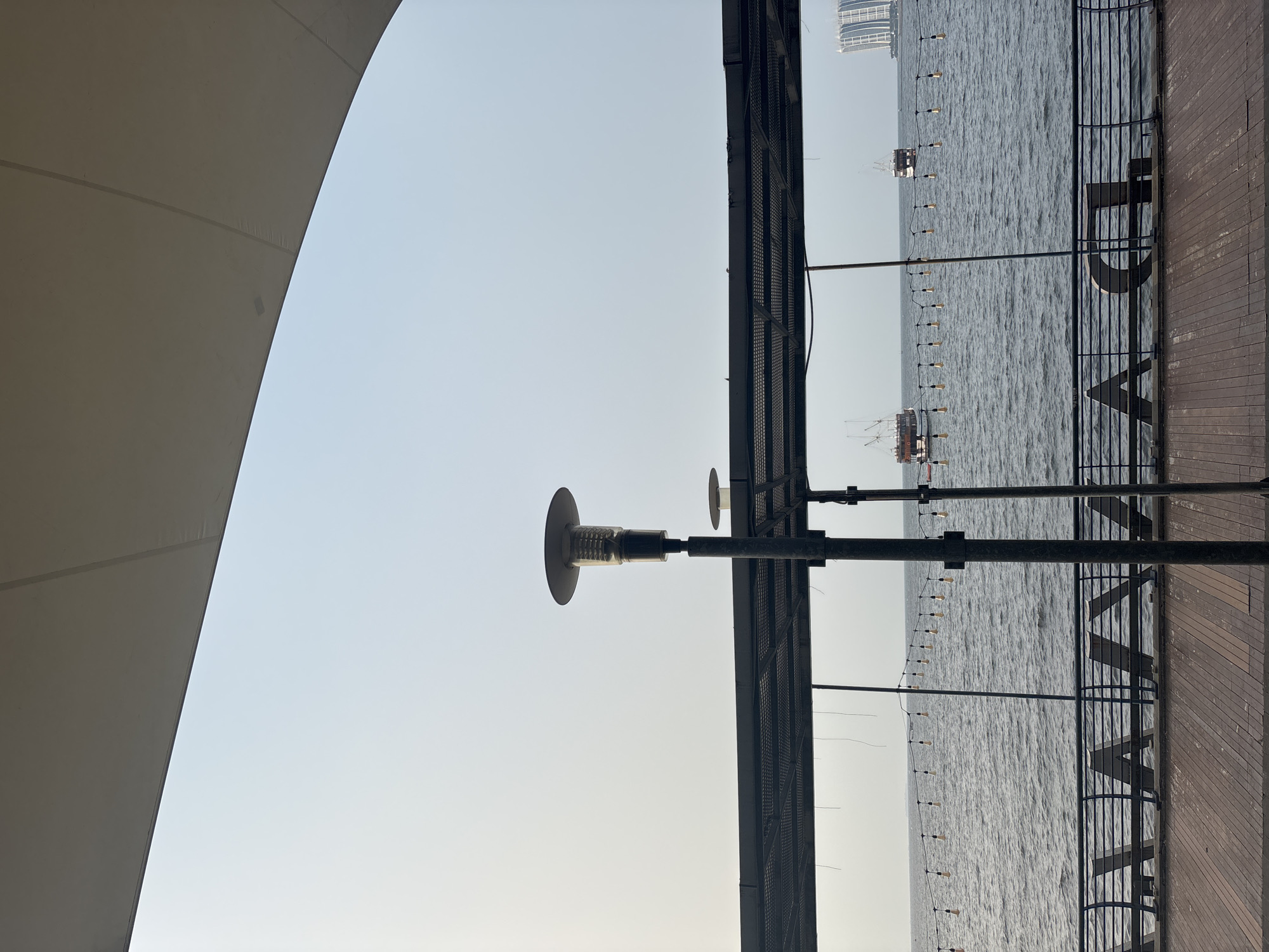 The image depicts an outdoor seating area overlooking a body of water, likely the Java Sea in North Jakarta, Indonesia. In the foreground, a wooden deck leads to a metal railing with decorative letter cutouts spelling out "LAGA" (Indonesian for "play" or "game"). Beyond the railing, a dark metal grate structure forms a canopy overhead, supported by posts. A tall, modern lamppost with a distinctive cap stands prominently on the right, with a smaller, similar lamppost visible behind it. A string of decorative bulbs stretches across the water, partially obscured by the canopy structure. The water itself is a calm, gray expanse, dotted with three boats: two traditional-looking vessels with multiple masts and a more modern structure to the far right. In the distance, several tall, modern buildings rise against a hazy, pale blue sky. The overall impression is one of a relaxed waterfront establishment.