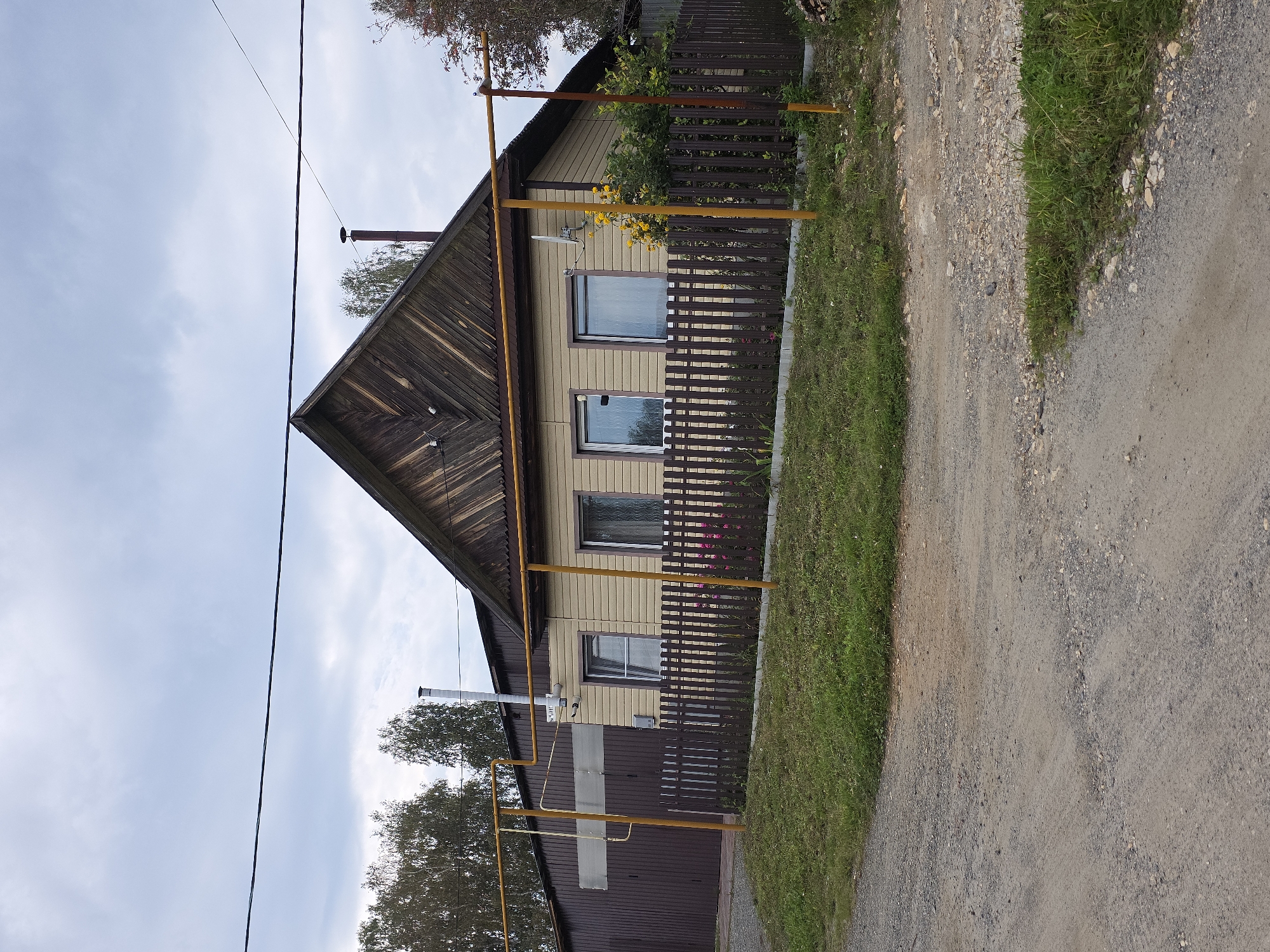 In the city of Magnitka, Russia, an exterior shot captures a rural dwelling with a brown-paneled garage to the left. The house itself features light-colored siding and a dark wooden, gabled roof. A dark wooden fence encloses a small yard with green grass and some flowering plants. Yellow gas pipes run horizontally across the front of the property, supported by vertical posts. A white chimney extends from the roof. The scene is set against an overcast sky, with trees visible in the background. A dirt road is in the foreground.