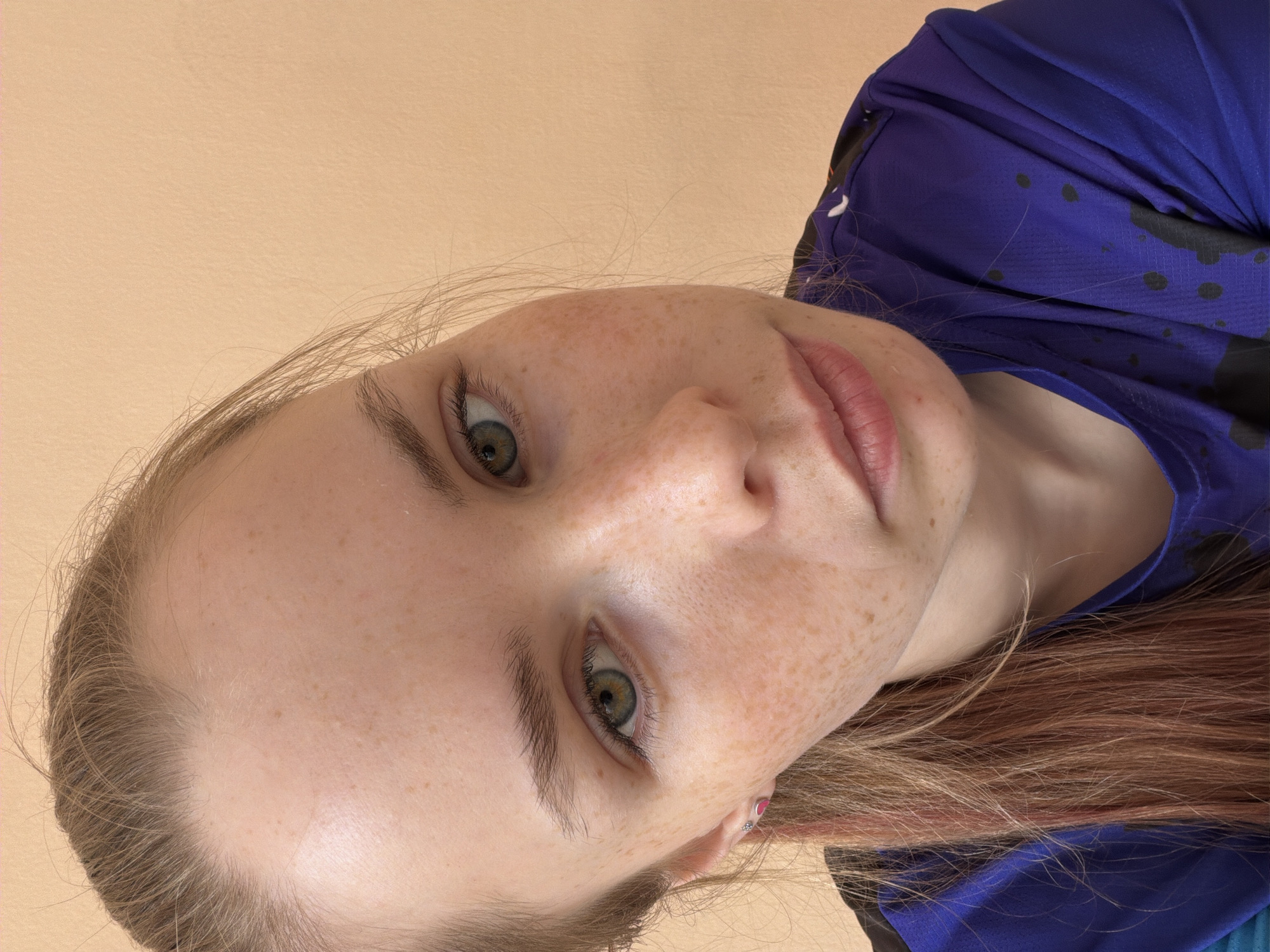 This is a close-up portrait of a young woman, captured from a low angle and significantly rotated counter-clockwise. Her face dominates the frame, with her head tilted and her eyes looking directly towards the viewer. She has fair skin, generously dotted with light brown freckles across her nose, cheeks, and forehead. Her eyes are a striking green or hazel, featuring distinct pupils and irises, complemented by visible eyelashes and well-defined, dark eyebrows. Her lips are naturally pink, slightly parted.
Her hair, a blend of light brown and blonde with some reddish undertones, frames her face and is visible on both sides of her head, extending towards the bottom right of the image. She is wearing a blue garment, possibly a casual top or sports attire, which features an abstract pattern of irregular dark, splattered shapes. A small, pink heart-shaped earring is visible in her right ear (on the left side of the image as rotated). The background is a plain, solid light peach or beige wall, offering a simple and uncluttered backdrop. The woman's expression is neutral and direct. No specific activity beyond posing for the camera is evident, and there is no visible text in the image. While the location is Berezniki, Russia, no visual cues within the photo confirm this.