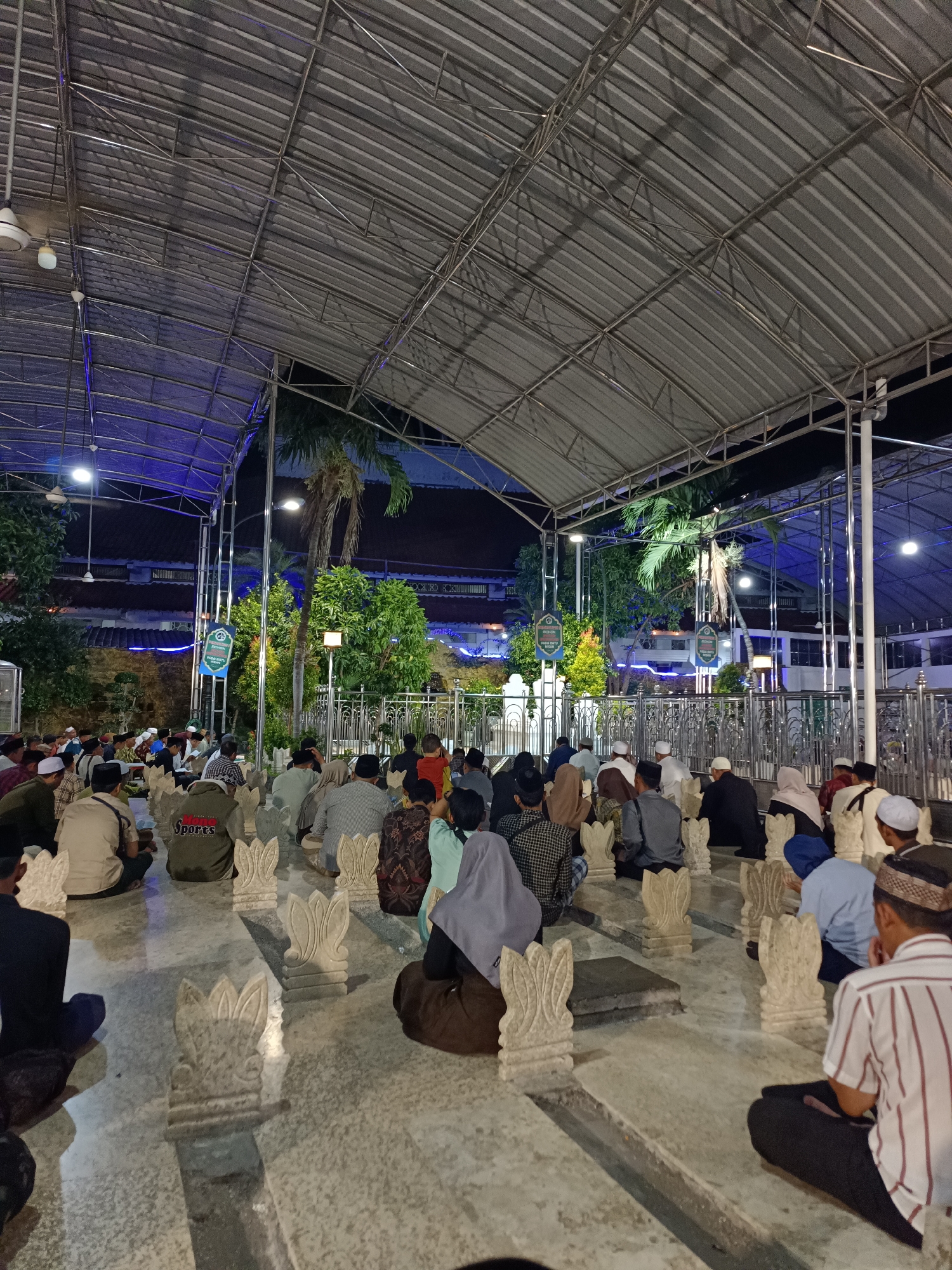 The image depicts a nighttime scene at a graveyard, likely in Surabaya, Indonesia, given the prompt. A large group of people, predominantly men, are seated on the ground in rows, facing a central area. They are dressed in casual attire, with many wearing traditional Islamic headwear such as peci or caps. Some individuals appear to be engaged in prayer or quiet contemplation.
The graveyard itself is covered by a large, metallic, corrugated roof supported by metal beams, suggesting an outdoor but sheltered area. The floor is paved with light-colored tiles, and there are numerous small, ornate headstones or grave markers scattered throughout the space. Decorative floral motifs are visible on some of these markers.
In the background, there are buildings with tiled roofs and some greenery, including palm trees, indicating a tropical or subtropical environment. Artificial lighting, including blue LED strips and white spotlights, illuminates the area, creating a somewhat ethereal atmosphere. Two blue signs with text in an unfamiliar script are visible, possibly indicating directions or information about the location. The overall impression is one of a communal gathering for religious observance or remembrance at a cemetery.