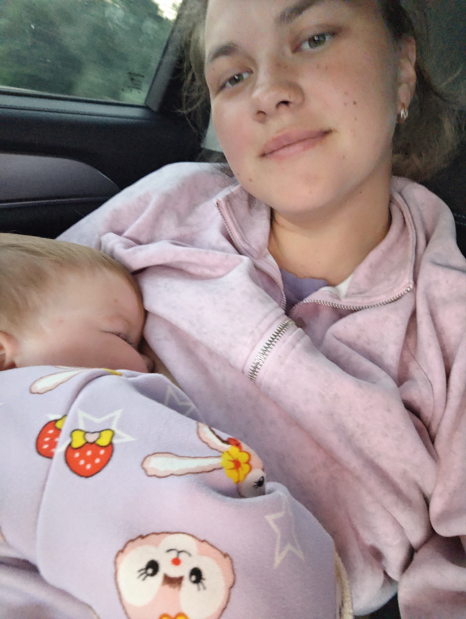 A young woman with fair skin and light brown hair is holding a baby. She is wearing a light pink zip-up jacket. The baby is swaddled in a light purple blanket with a print of strawberries and owls. They are likely in a car due to the presence of a car window and interior. It is daytime, based on the outdoor light. The location is not discernible from the image; there is no evidence to suggest the city of Noviy, Russia.
