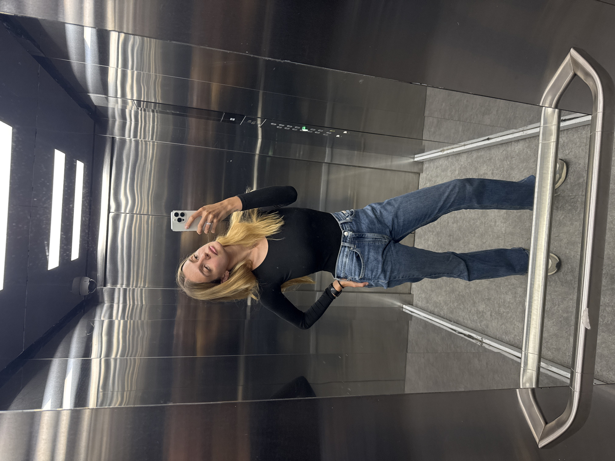 A young blonde woman is taking a selfie in an elevator. She is wearing a long-sleeved black shirt and blue jeans. Her phone is white with a silver frame. The elevator interior is predominantly stainless steel with horizontal lines and a row of lighted floor buttons. There's a safety bar on the right side of the elevator. The floor is gray. There is a security camera installed in the top left corner. The location is Unknown, Unknown.