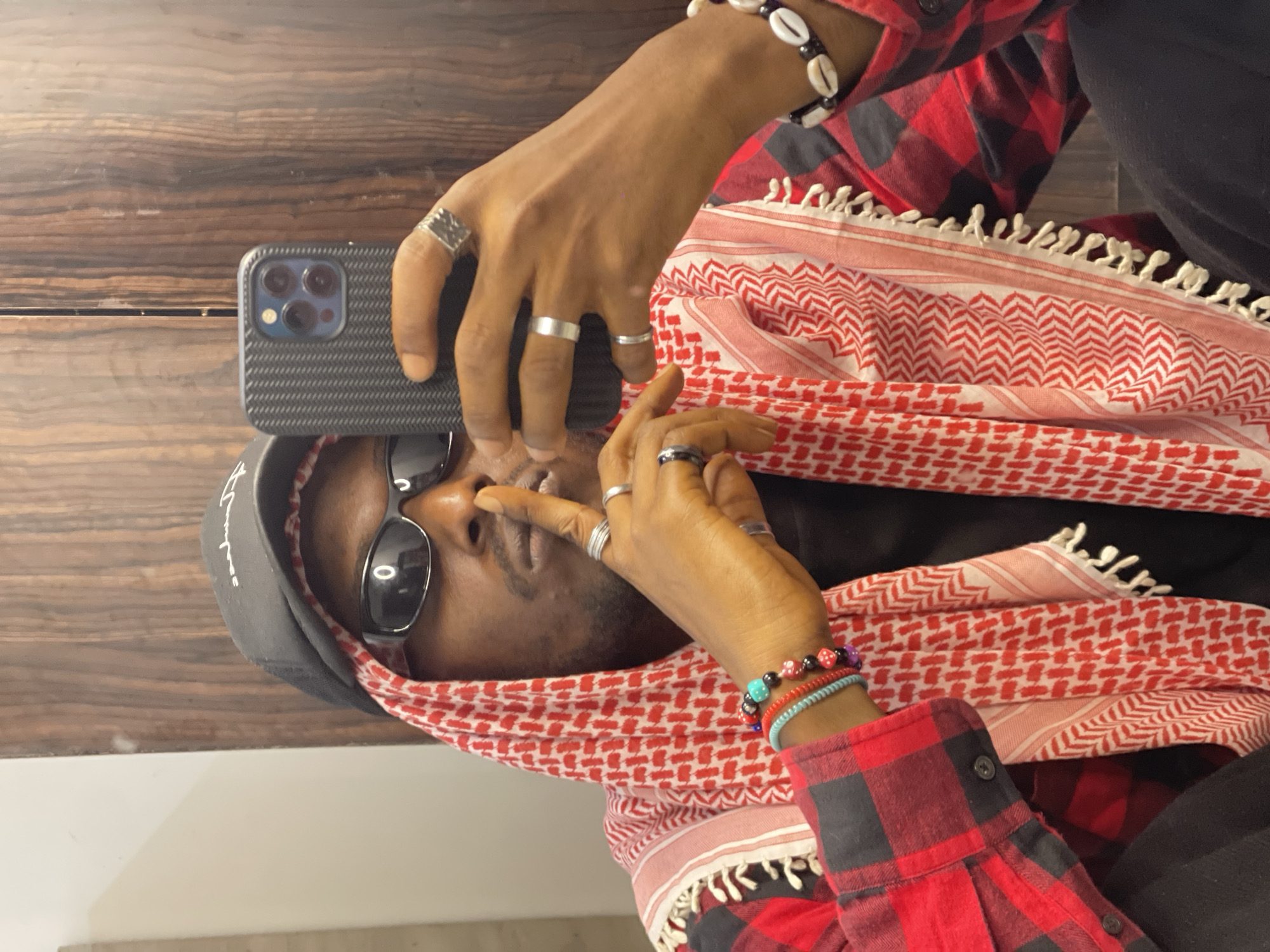 A selfie taken in a bathroom shows a Black man wearing a black baseball cap, black sunglasses, a red and black plaid shirt, and a red and white keffiyeh. He is holding a smartphone in his right hand, taking a picture of himself in the mirror. His left hand is raised to his lips in a "shushing" gesture. He is wearing several rings and bracelets. The background appears to be a wooden cabinet door. The image was taken in Abuja, Nigeria.
