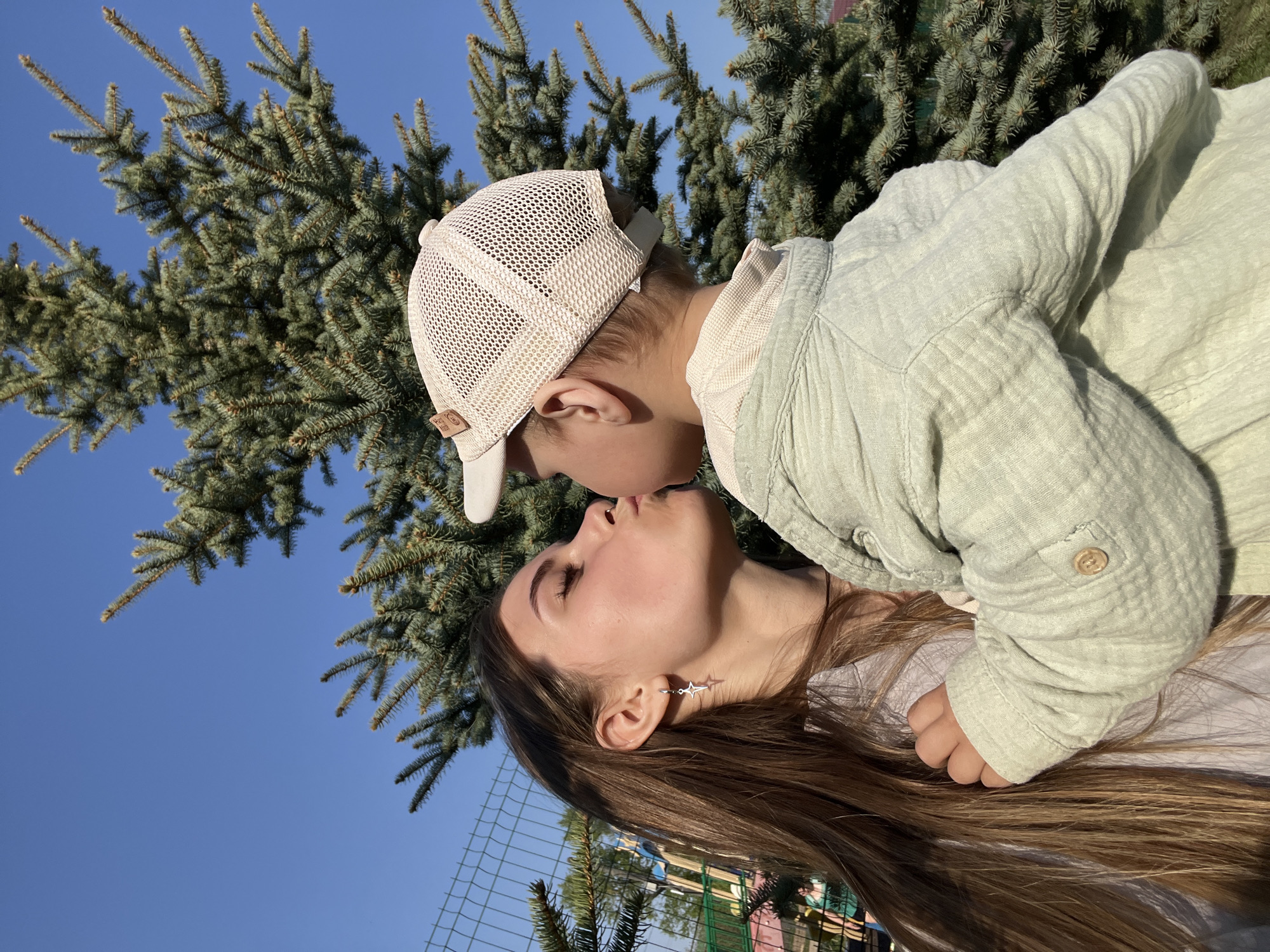 A fair-skinned woman with long brown hair and a star-shaped earring is being kissed by a young child wearing a light-colored baseball cap and light green shirt. They are outside, near a tree with blue-green needles, under a clear blue sky. A wire fence is visible in the background. The location is likely near Фоки, Russia based on the location query.