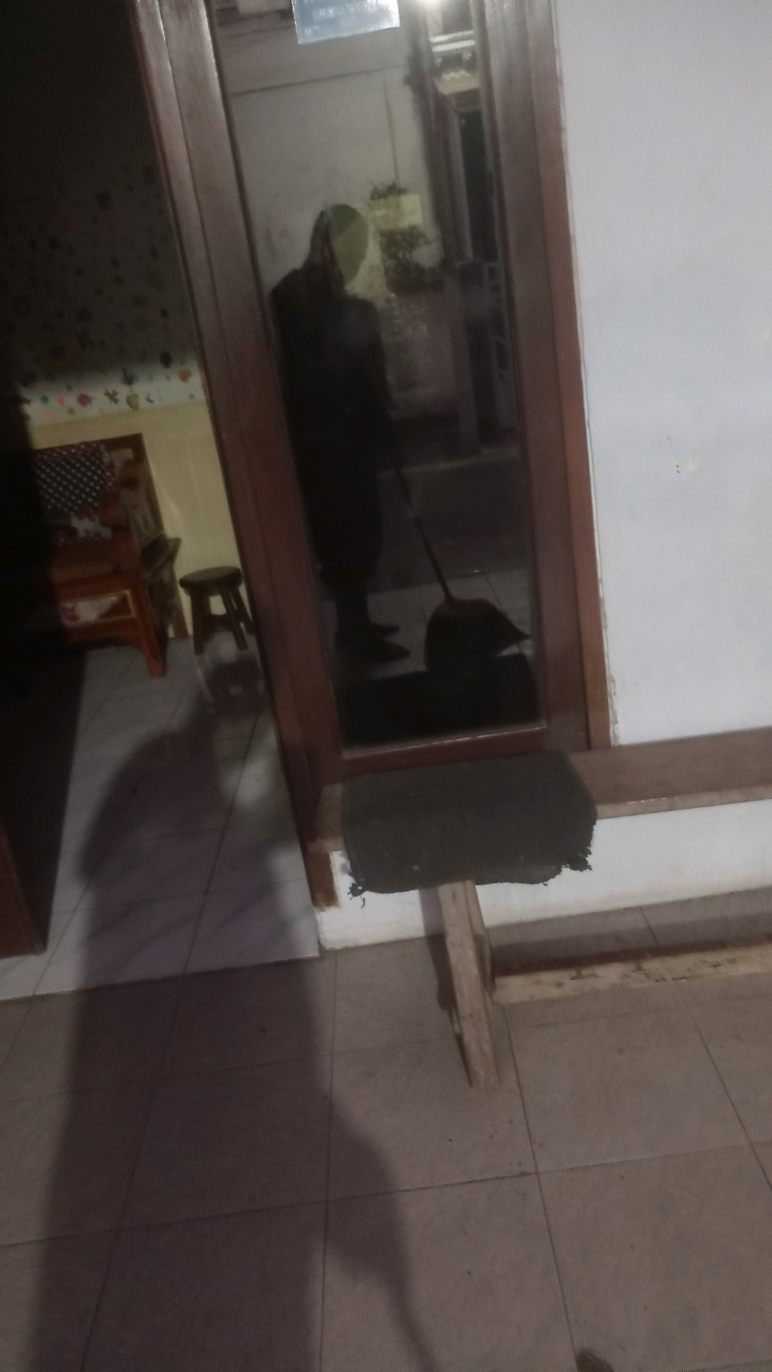 The image is a photograph taken at night or in low light conditions, likely in Mulyoagung, Indonesia, given the context provided. The scene appears to be an interior space, possibly a home, with tiled flooring.
In the foreground, the tiled floor is visible, with a large shadow cast across it, suggesting the presence of a person or object outside the frame or not fully captured. To the right, a wooden plank or shelf is positioned horizontally, supported by wooden legs. A dark-colored mat is placed on this plank.
A doorway or large window with a wooden frame occupies the center and left side of the image. Through the glass, a reflection of a person can be seen. This individual is wearing dark clothing and appears to be holding a broom and dustpan, engaged in sweeping. The reflection also shows some interior elements like a wall with decorative elements or wallpaper and a piece of furniture.
To the far left, a portion of another piece of furniture, possibly a chair or table, is visible with a patterned cushion. A small wooden stool is also present in this area. The overall atmosphere is dimly lit, and the image quality is somewhat grainy, consistent with low-light photography. The scene suggests a domestic setting where cleaning is in progress.