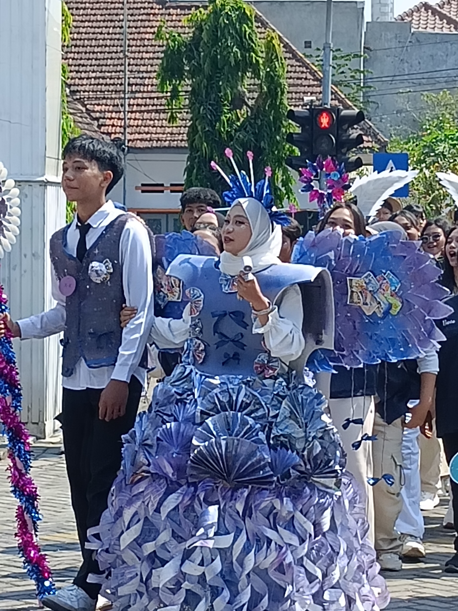 In Kediri City, Indonesia, a parade is taking place on a sunny day. A young woman wearing a blue and white hijab and a creatively designed blue costume with intricate folded paper embellishments is the central figure. She holds a microphone and appears to be addressing the crowd. To her left, a young man in a white shirt, black tie, and a decorated grey vest walks alongside her, holding a purple and pink tinsel garland. Behind them, other participants are visible, some also wearing elaborate costumes, including large blue fan-like structures adorned with what appear to be printed images. In the background, traffic lights are visible, with the red "don't walk" signal illuminated. Buildings with tiled roofs and green trees line the street, suggesting an urban or semi-urban setting. The overall scene depicts a lively community event or parade.