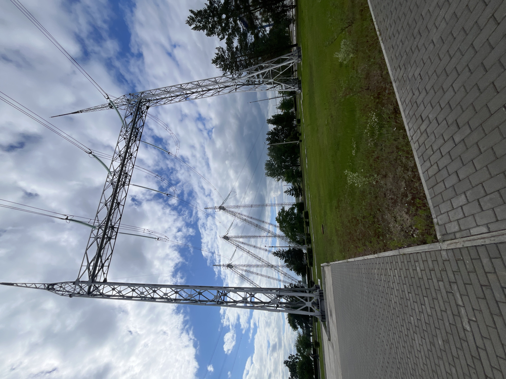 A wide-angle, low-angle shot captures a power substation under a partly cloudy sky, with a paved walkway in the foreground. The primary focus is a towering metal electricity pylon, its lattice structure supporting thick power lines that stretch across the frame. In the background, more pylons and lines are visible, extending towards a line of trees and a building. The ground is covered in green grass, and the paved walkway is made of gray interlocking bricks. The scene appears to be a part of the infrastructure of Chaikovsky, Russia. There are no visible people in the image.