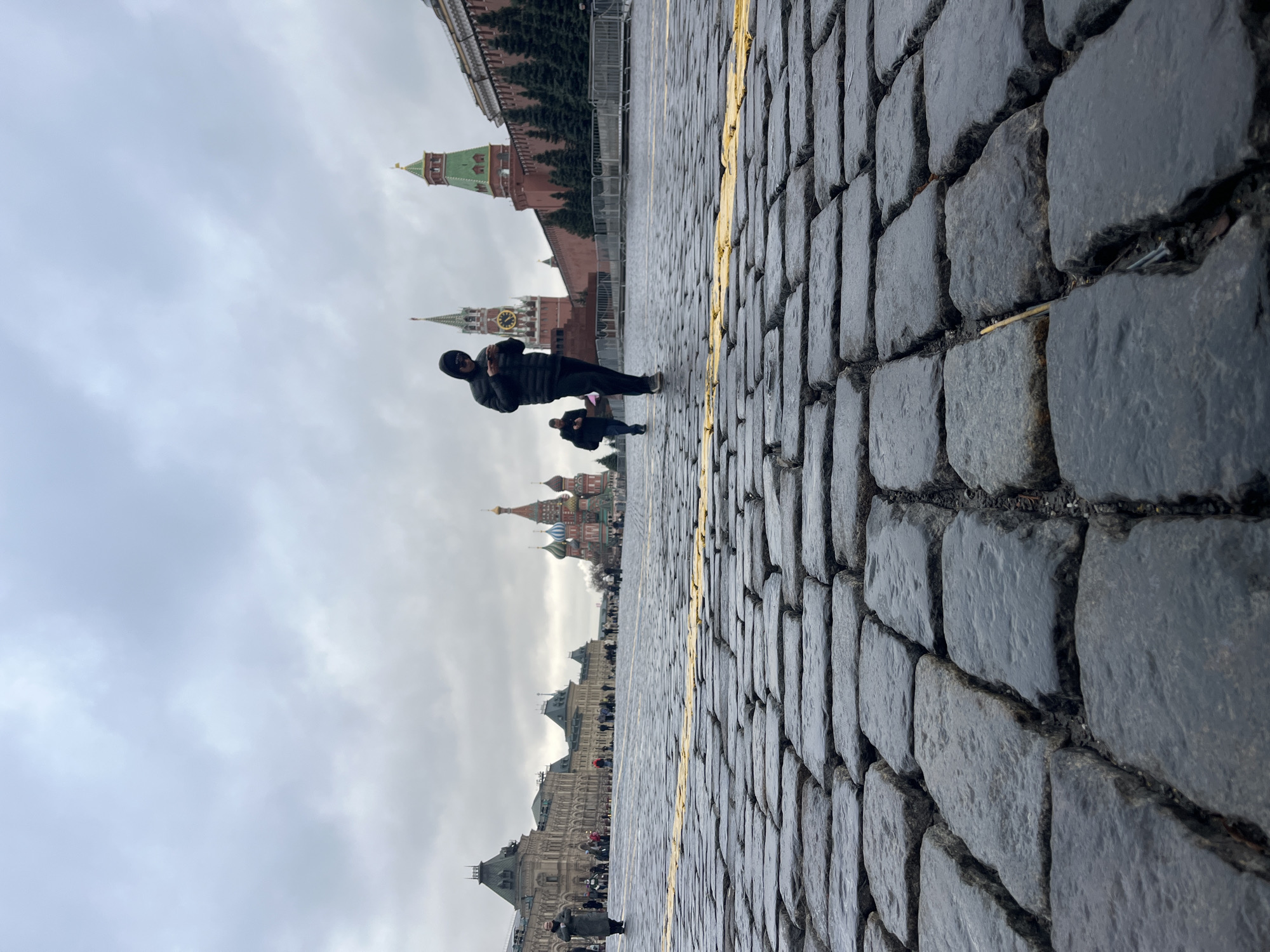 The image depicts Red Square in Moscow, Russia, under an overcast sky. The foreground is dominated by a cobblestone pavement with a painted yellow line running along it. In the midground, there are two people: one is standing and appears to be looking at their phone, wearing a black jacket and a mask covering their face; the other person is crouched down. In the background, the image features the various landmarks of Red Square, including the Kremlin walls and towers, and St. Basil's Cathedral. Other people are walking along the square.