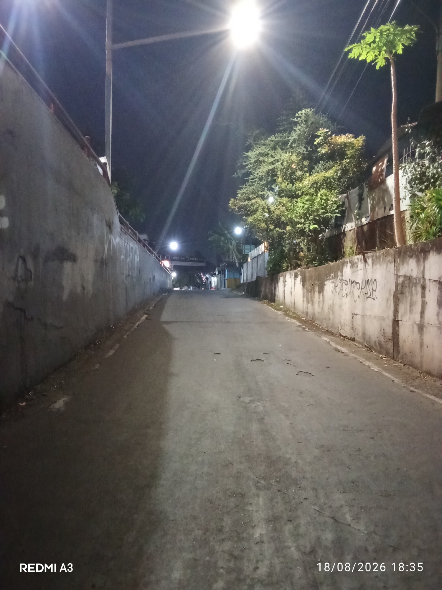 An outdoor, eye-level, vertical-format, nighttime photograph captures a deserted, narrow, paved road in Purbayan, Indonesia. The road slopes upward into the distance, illuminated by a bright overhead street lamp casting a strong glare to the left. To the left of the road is a tall, gray concrete wall with some graffiti. To the right of the road is a dense cluster of green trees and foliage, with a thin, tall tree with light green leaves standing prominently in the foreground. Beyond the trees, structures and more foliage are visible. In the far distance, two small figures are barely discernible. The road surface shows some wear and tear, with small cracks and debris. The bottom left of the image displays the text "REDMI A3," and the bottom right displays "18/08/2026 18:35."