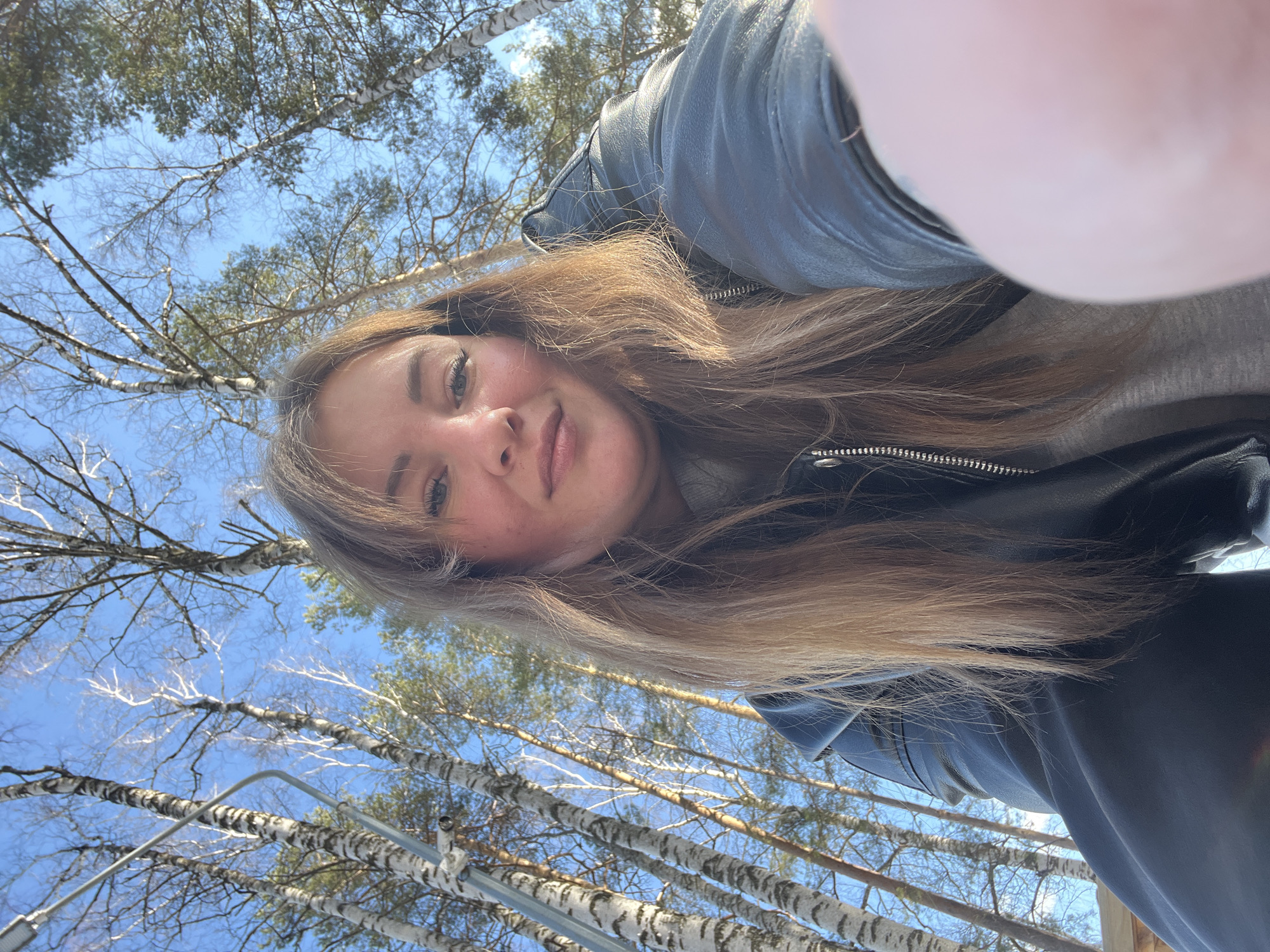 A young woman takes a selfie outdoors. She wears a gray shirt under a black leather jacket. Her long, light-brown hair falls across her chest. Behind her are tall, slender birch trees with white bark, reaching into a blue sky with few clouds. Based on the trees and sky, it is likely the photo was taken in a park or wooded area in Chaikovsky, Russia, considering the query specifies the location. A portion of her finger covers part of the upper right side of the frame.