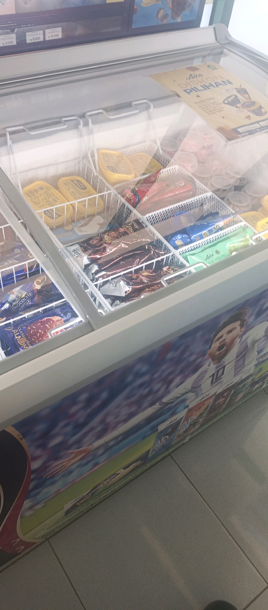 The image shows a freezer full of ice cream. The freezer is decorated with a picture of Lionel Messi, a famous soccer player. There are several brands of ice cream in the freezer, including Aice, which is a popular brand in Indonesia. The freezer is located in a store in Lamongan, Indonesia, a city known for its culinary scene and agricultural products. The ice cream selection appears to be a mix of popular flavors and brands, suggesting it is a well-stocked freezer in a convenience store or supermarket.