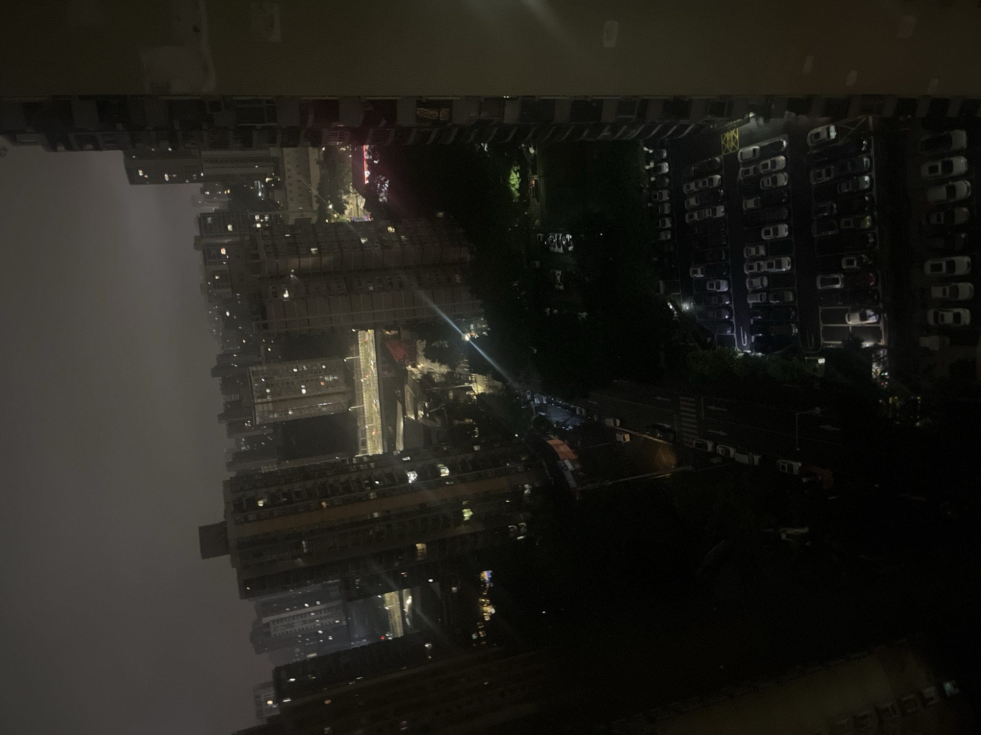 The image depicts a nighttime urban landscape in Jiulongpo District, China, viewed from a high vantage point, likely a tall building. The sky is heavily overcast and dark, with a muted ambient glow from widespread city lights.
The scene is dominated by numerous high-rise residential buildings, densely packed across the frame, with many windows illuminated, indicating occupancy. An elevated multi-lane highway or overpass with visible vehicle traffic cuts across the mid-ground, showing lines of light from moving cars. Below the highway, a network of darker local roads is discernible, also with vehicle traffic and streetlights.
Several parking lots are visible throughout the lower and mid-ground. The most prominent is in the lower right, brightly lit and filled with numerous parked cars, clearly showing white parking lines and yellow directional markings. Other, less illuminated parking areas are also present. Patches of dark green foliage from trees are interspersed between buildings and along roads, suggesting small park areas or planted strips.
No individuals are discernible, and specific actions or interactions beyond vehicle movement on roads are not visible. The primary subjects are the urban infrastructure, residential buildings, vehicles, and the interplay of artificial light against the dark, cloudy night.