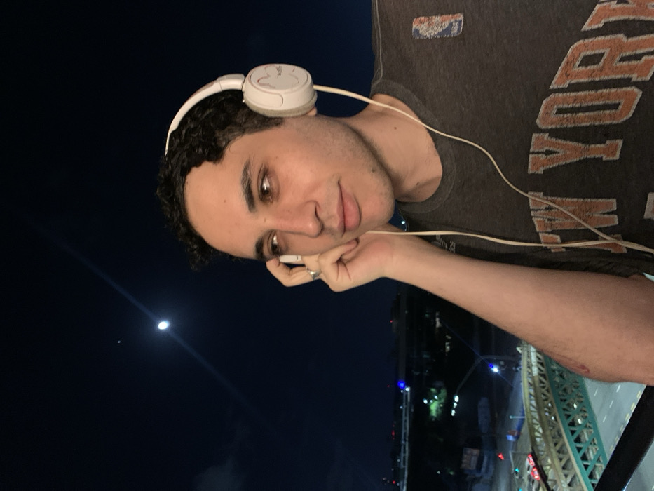 A person is facing the camera at night, wearing headphones and a New York t-shirt. The person has curly dark hair, light skin, and brown eyes. The person is propping his head up with his hand. The background shows a dark night sky with a bright moon. City lights and a bridge structure are visible in the distance.