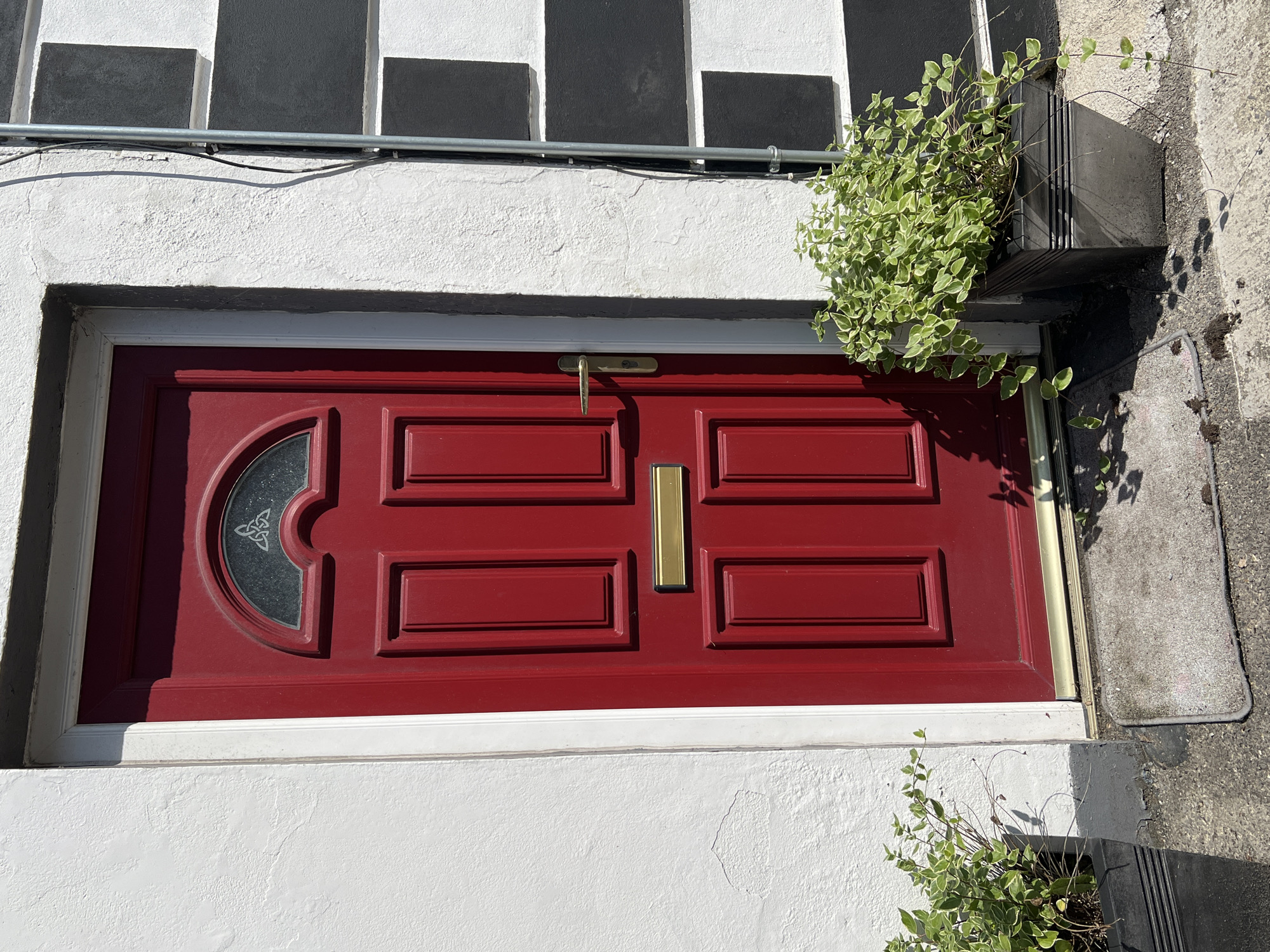 A front door in the city of Donegal, Ireland, is shown. The door is a bright red, rectangular object with a semi-circular glass panel at the top featuring a Celtic knot design. It has a gold-colored mail slot and a matching gold-colored doorknob. The door is framed by a white trim and is set into a rough, white-painted wall. To the right of the door, the wall transitions to a black and white striped pattern, with thick horizontal stripes. A metal conduit runs vertically down the wall. In front of the door, on the left side, is a dark rectangular planter containing a lush green plant with trailing vines. Another similar planter with a matching plant is positioned to the right of the door. A small, gray doormat lies on the concrete pathway in front of the door. The ground is a textured concrete. The scene is illuminated by bright sunlight, casting shadows from the plants and door.