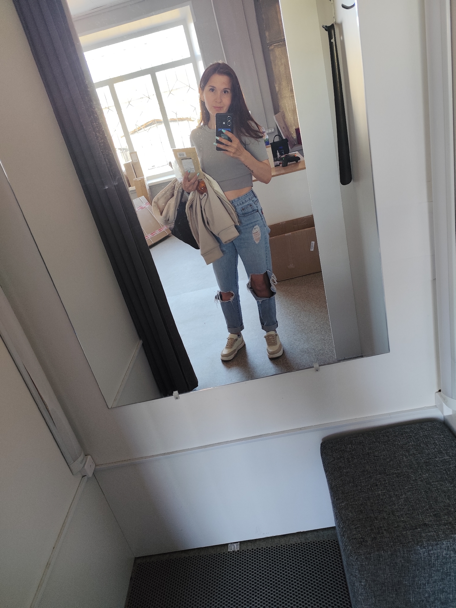 A full-length shot depicts a woman taking a mirror selfie in a changing room. The woman is wearing a gray crop top, ripped jeans, and sneakers. She holds a smartphone in her right hand and a light-colored jacket and a cardboard package in her left hand. The changing room is small, with a gray curtain on the left and a gray upholstered seat on the bottom right. The mirror frames the woman and shows a glimpse of the outside through a window in the background, where boxes are visible. A black shoehorn is attached to the right wall. The location is not discernible as Фоки, Russia, based on the given information.