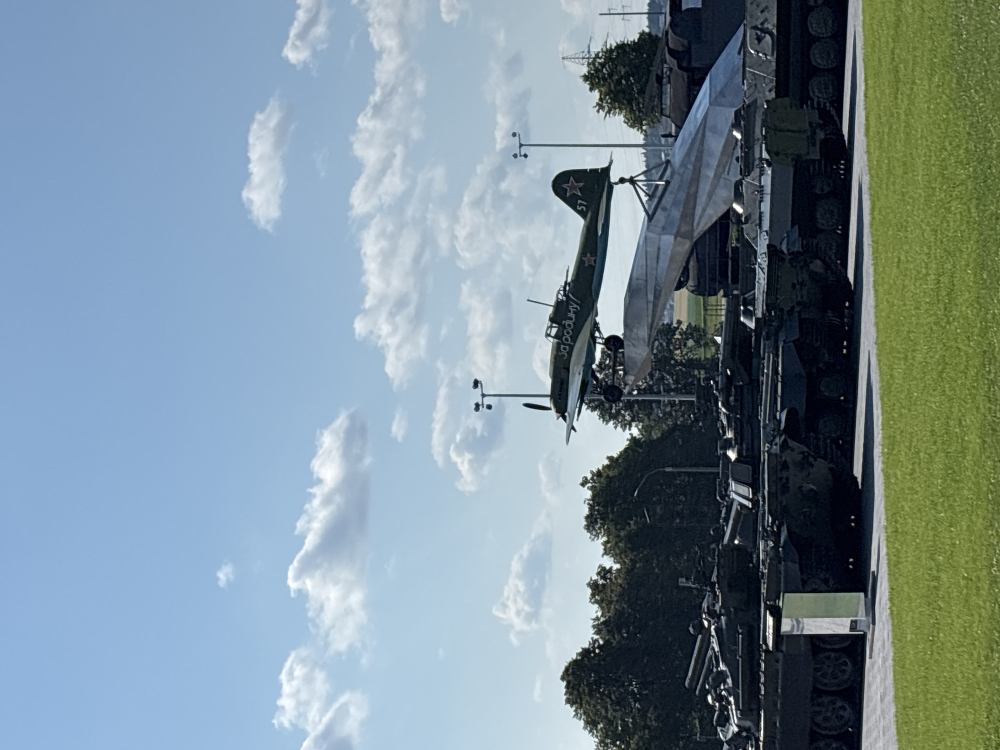 The image depicts an outdoor exhibit of military vehicles under a partly cloudy sky. In the foreground, a line of dark-colored tanks is parked on a paved surface, with a section of green grass to the left. Behind the tanks, a large, abstract metal sculpture is partially visible, supporting a green airplane with red stars on its wings. The airplane has Cyrillic lettering on its fuselage. Trees form a dense line across the middle ground, and a power line and transmission tower can be seen in the distance to the right. The scene appears to be a memorial or museum dedicated to military history, likely located in the Ozyaritska-Slabadski rural council of Belarus, given the military hardware and potential inscription on the plane. There are no people visible in the image.