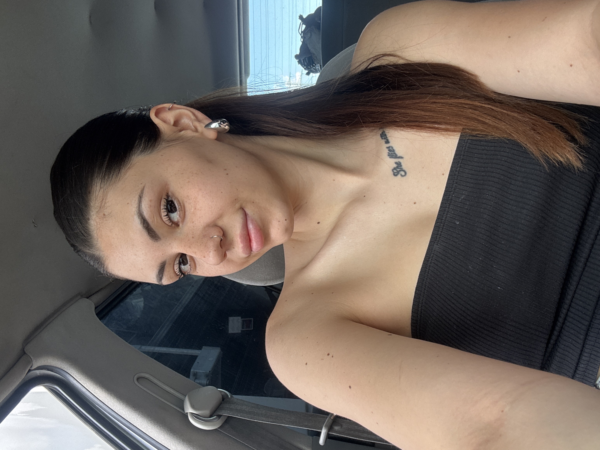 The image displays a young adult female taking a selfie from the passenger seat of an automobile. She has dark brown hair, pulled back from her face and cascading over her right shoulder. Her complexion features numerous freckles across her nose and cheeks. She wears a small silver hoop nose ring on her right nostril. Her left earlobe holds a large, reflective silver teardrop-shaped earring, with a smaller silver hoop in her upper helix. She is dressed in a black, ribbed, strapless tube top. A tattoo with the partial text "She flies with" is visible on her upper left chest/collarbone area. Her right arm and shoulder have several moles, and faint linear marks are present on the inside of her right upper arm.
The car interior features gray upholstery on the B-pillar, seatbelt mechanism, and overhead lining. Blurred daylight is visible through the side and rear windows, showing generic outdoor scenery without specific identifiable landmarks. The rear window exhibits faint horizontal defroster lines. No elements within the image provide discernible information regarding a specific city or country, including Sofia, Bulgaria.