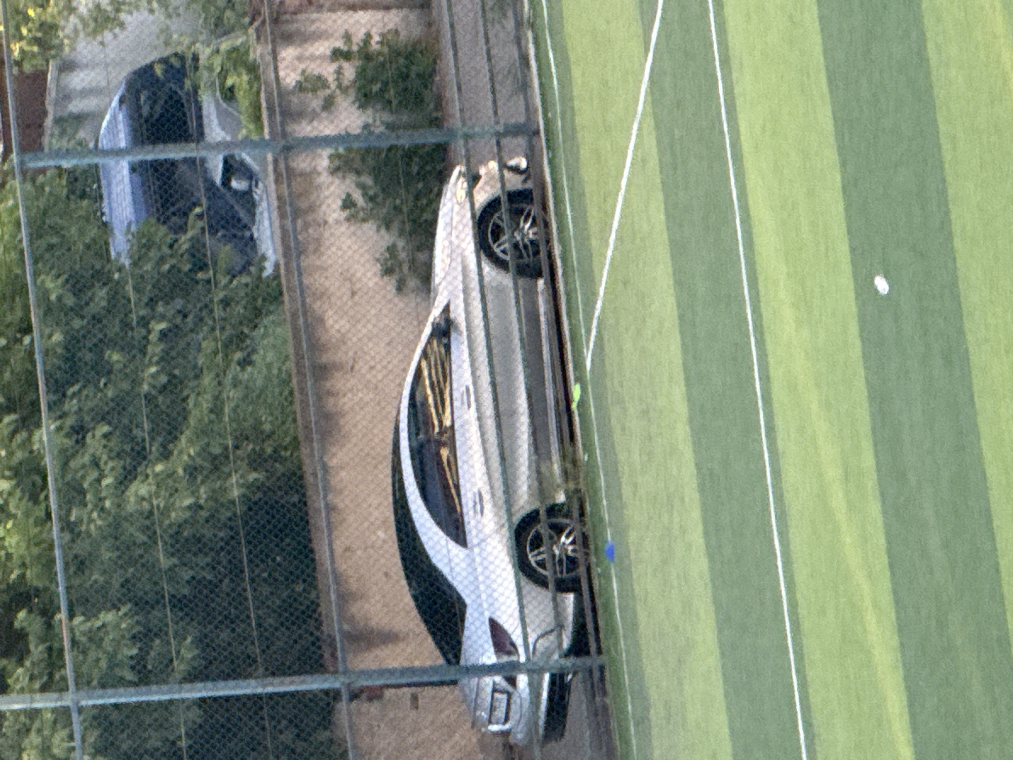 The image depicts a silver Mercedes-Benz sedan parked next to a chain-link fence, with a green and white striped surface resembling a tennis court in the foreground. Behind the fence, a dirt embankment is visible, topped by another fence and then lush green foliage. In the background, through the foliage and a fence, parts of two vehicles, possibly SUVs or vans, can be seen. One is a dark blue or black vehicle, and the other appears to be lighter in color. The lighting suggests it is daytime. The location is identified as Tbilisi, Georgia.