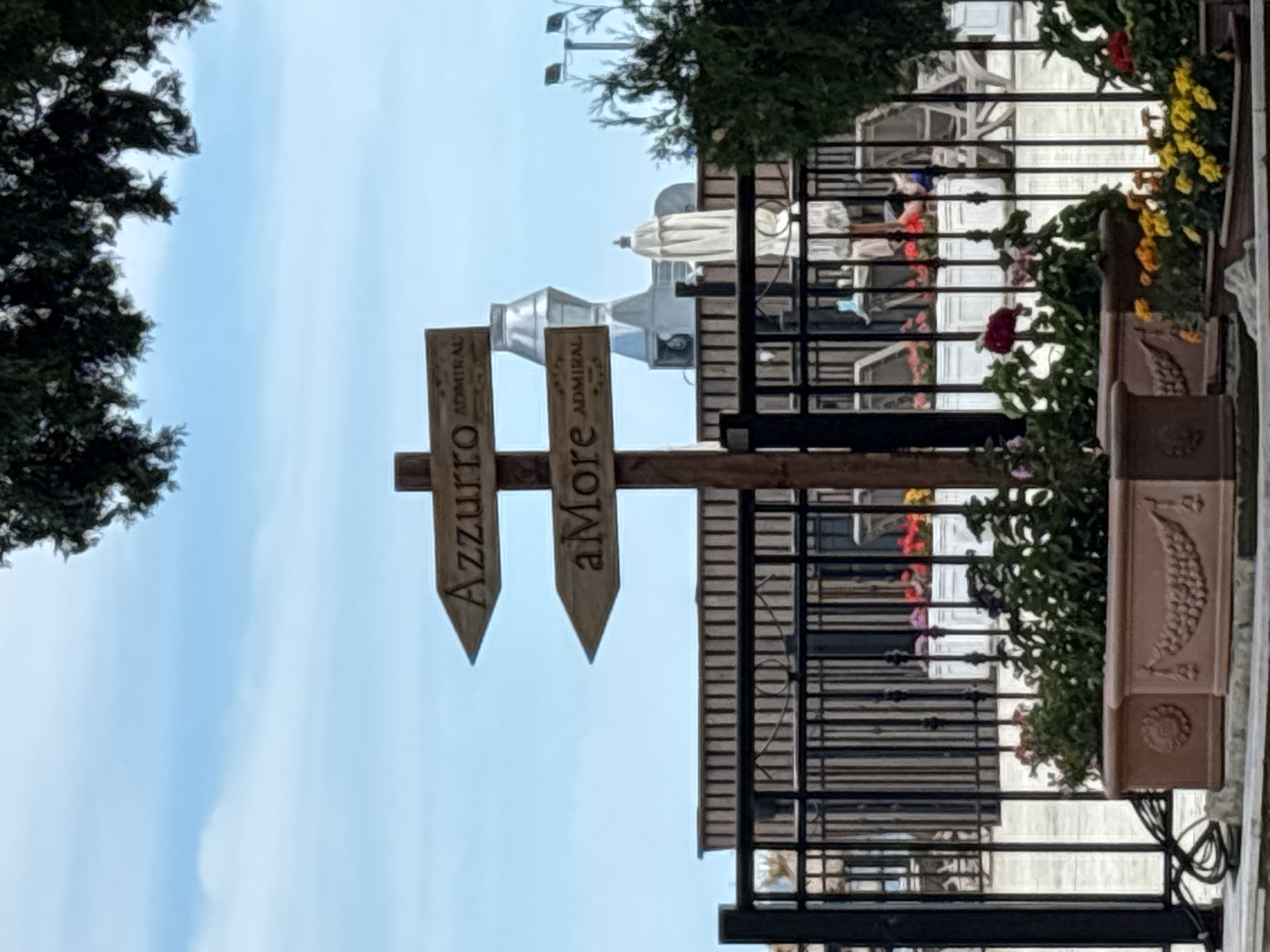 The image depicts a directional wooden sign with two arrows pointing left. The top arrow reads "Azzurro ADMIRAL," and the bottom arrow reads "aMore ADMIRAL." The sign is planted in the ground in front of a black metal fence. Behind the fence, there is a row of planters with colorful flowers, a light-colored building with a dark horizontal striped facade, and some outdoor furniture including chairs and tables. The sky is blue with some white clouds. A large tree with dark green leaves partially covers the top of the image. This sign is likely located at the Admiral Hotel in Varna, Bulgaria, as indicated by the text on the sign.
