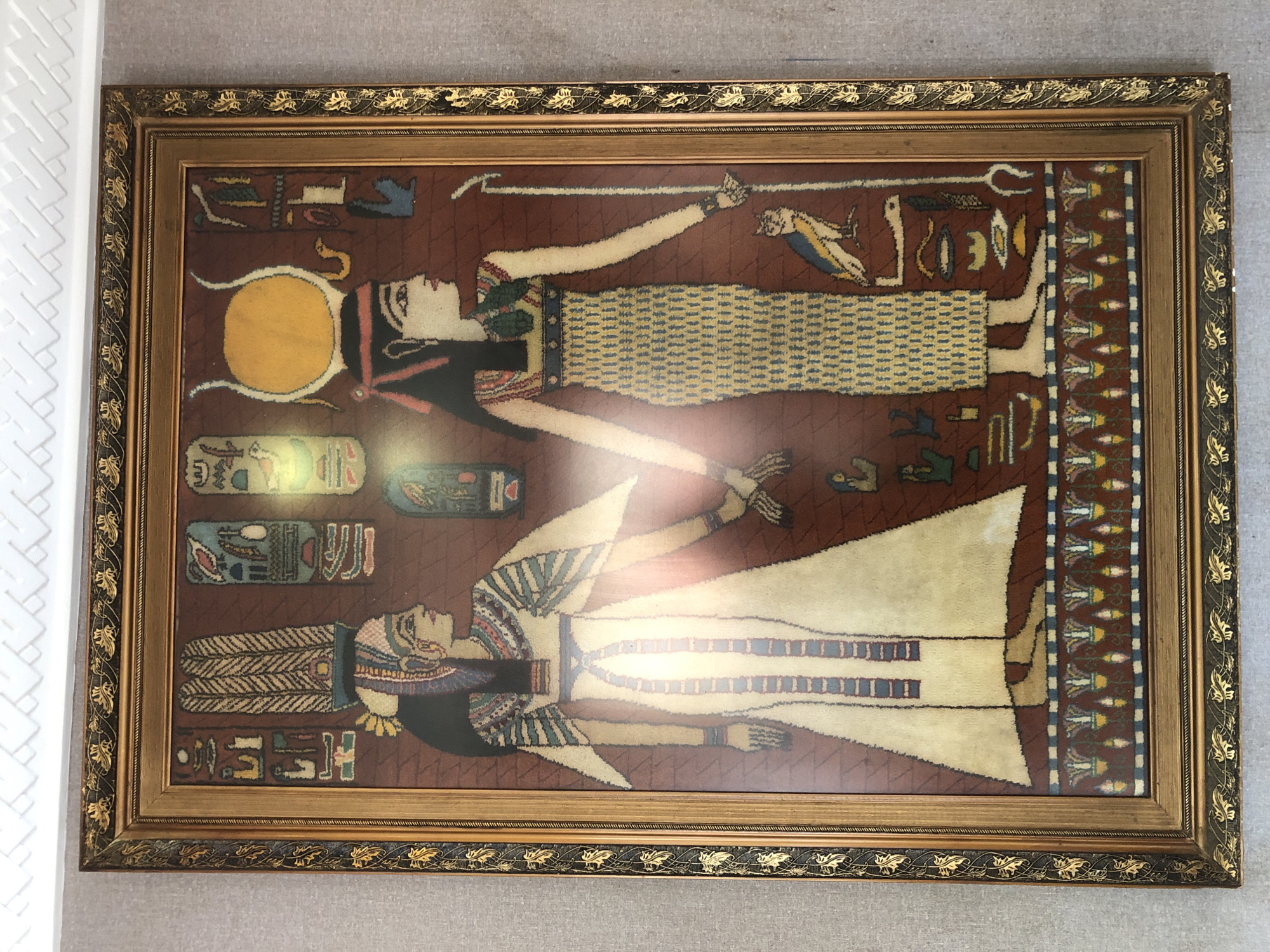 A framed Egyptian tapestry hangs on a neutral-colored wall. The frame is ornate, with gold leaf and a dark inner border. The tapestry depicts two stylized figures in profile. The upper figure wears a yellow patterned garment, while the lower figure wears white. Both figures have dark hair and Egyptian headdresses. Hieroglyphs and other symbols are arranged around the figures. The scene seems to be indoors. The location is in the city of Medan, Indonesia.