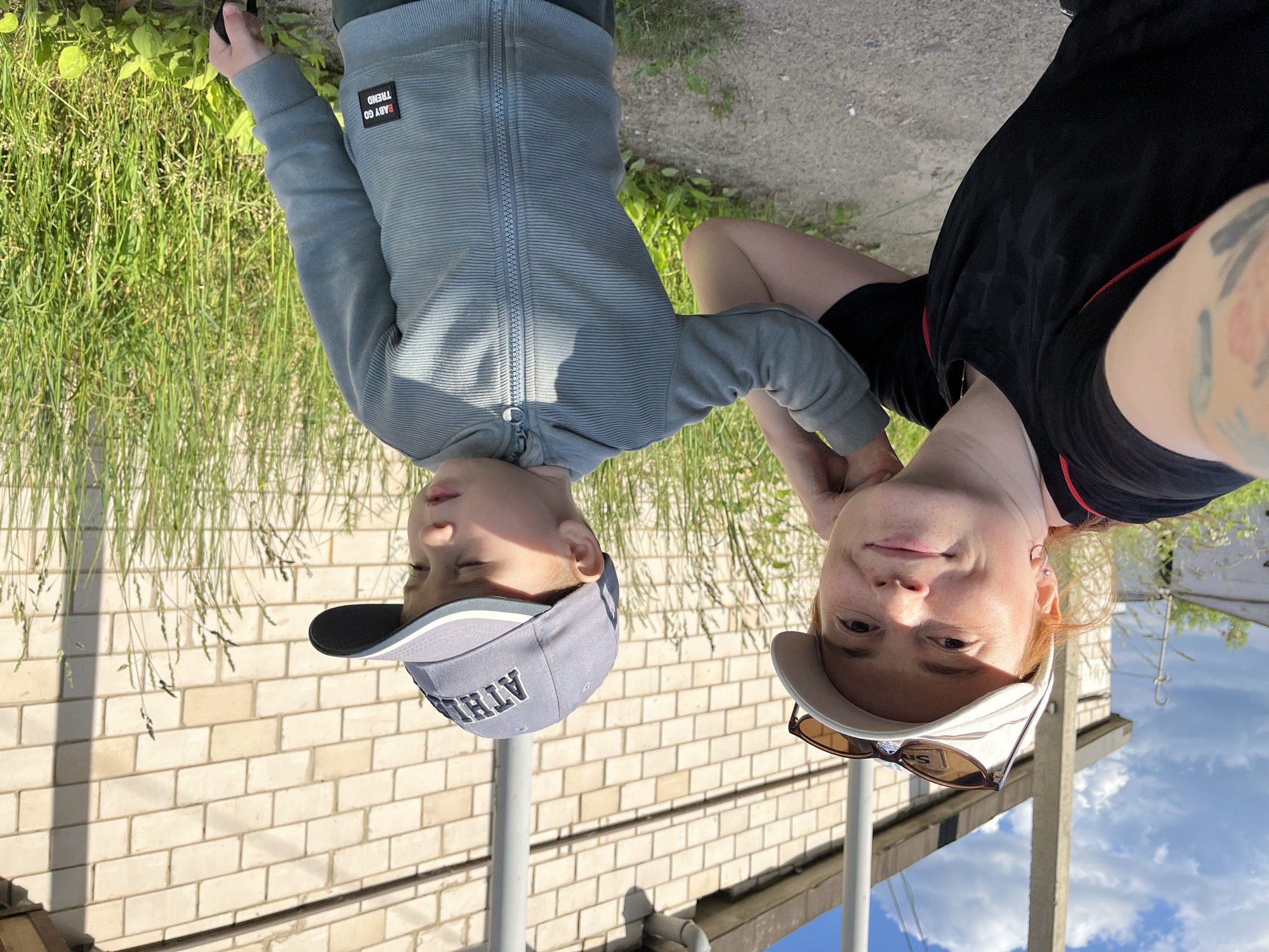 A close-up, upside-down selfie shows two people in an outdoor setting with a brick wall and grass in the background. The person in the foreground on the right, with orange hair and fair skin, is wearing a black t-shirt and white cap with sunglasses perched on the brim. They have freckles and are wearing a silver hoop earring. The person on the left, a young boy, is wearing a grey zip-up hoodie and a grey baseball cap. The hoodie has a small black and red patch on the left chest with text that reads "BABY GO TREND". The boy's cap has a dark brim and the word "ATHL." is visible in white lettering. Both individuals appear to be looking slightly off-camera, with neutral expressions. The lighting suggests it is daytime, likely sunny given the shadows and bright sky visible in the upper right portion of the image. The background includes lush green grass and a light-colored brick wall, possibly part of a building. The overall scene suggests a casual, outdoor activity.