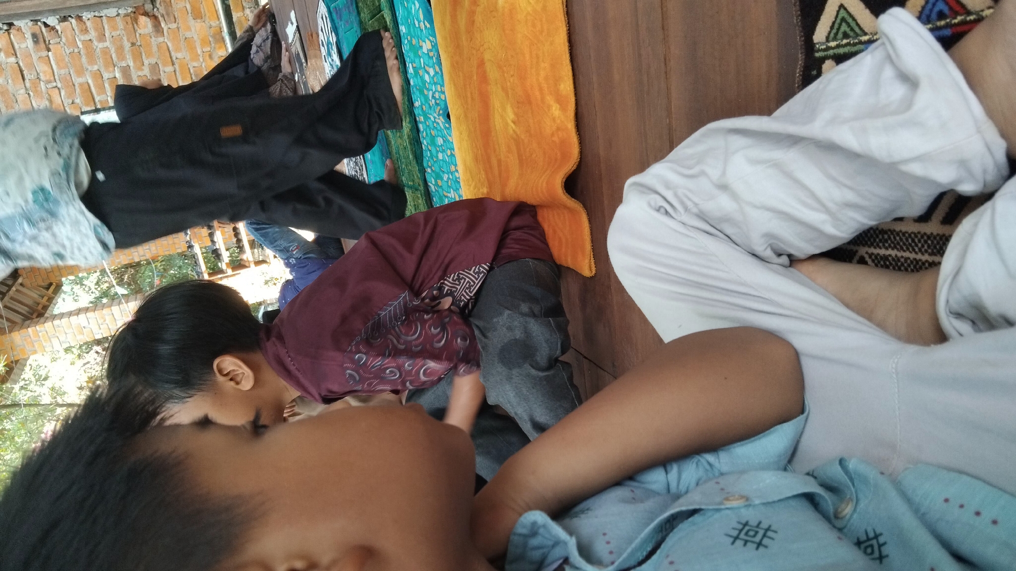 In Bangun Rejo, Indonesia, two young boys are seated on a wooden floor. One boy, in the foreground and to the left, wears a light blue shirt with a geometric pattern and white pants. His arm is bent, and his head is tilted downwards. The second boy, positioned behind and to the right of the first, wears a maroon shirt with intricate white and black patterns and dark pants. He is looking to his left. Between them, an orange mat is visible, and beyond them, several other people are present, some standing and some seated. A brick wall and a wooden roof structure are also part of the environment, suggesting an outdoor or semi-open setting.