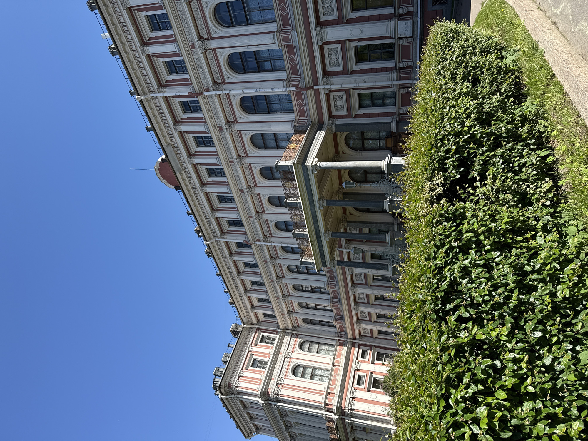 The image, originally rotated 90 degrees clockwise, captures a majestic, ornate building under a clear, bright blue sky, partially framed by a dense green hedge.
The grand, multi-story building, characteristic of Neoclassical or Baroque architecture, occupies the left and central parts of the frame. Its facade is a combination of off-white or cream, accentuated with red-brown decorative elements, including pilasters, cornices, and window surrounds. The building features numerous windows, many with elegant arched tops, and several balconies adorned with intricate dark metal railings. A prominent entrance or portico with classical columns is partially visible at a lower level, mostly obscured by the foreground foliage. Towards the building's roofline, a small, reddish dome and various ventilation structures are discernible.
A well-maintained, lush green hedge runs along the right side of the image, separating the building from a lighter green strip of grass and a paved path.
The sky, dominating the upper left portion of the photograph, is a vivid, cloudless blue, indicating a clear day. Bright sunlight illuminates the scene, casting distinct shadows that emphasize the architectural details. While there are small plaques on the building's facade, any text on them is too indistinct to read. The architectural style aligns with the provided location context of Saint Petersburg, Russia. The scene is static, with no people or specific activities taking place.