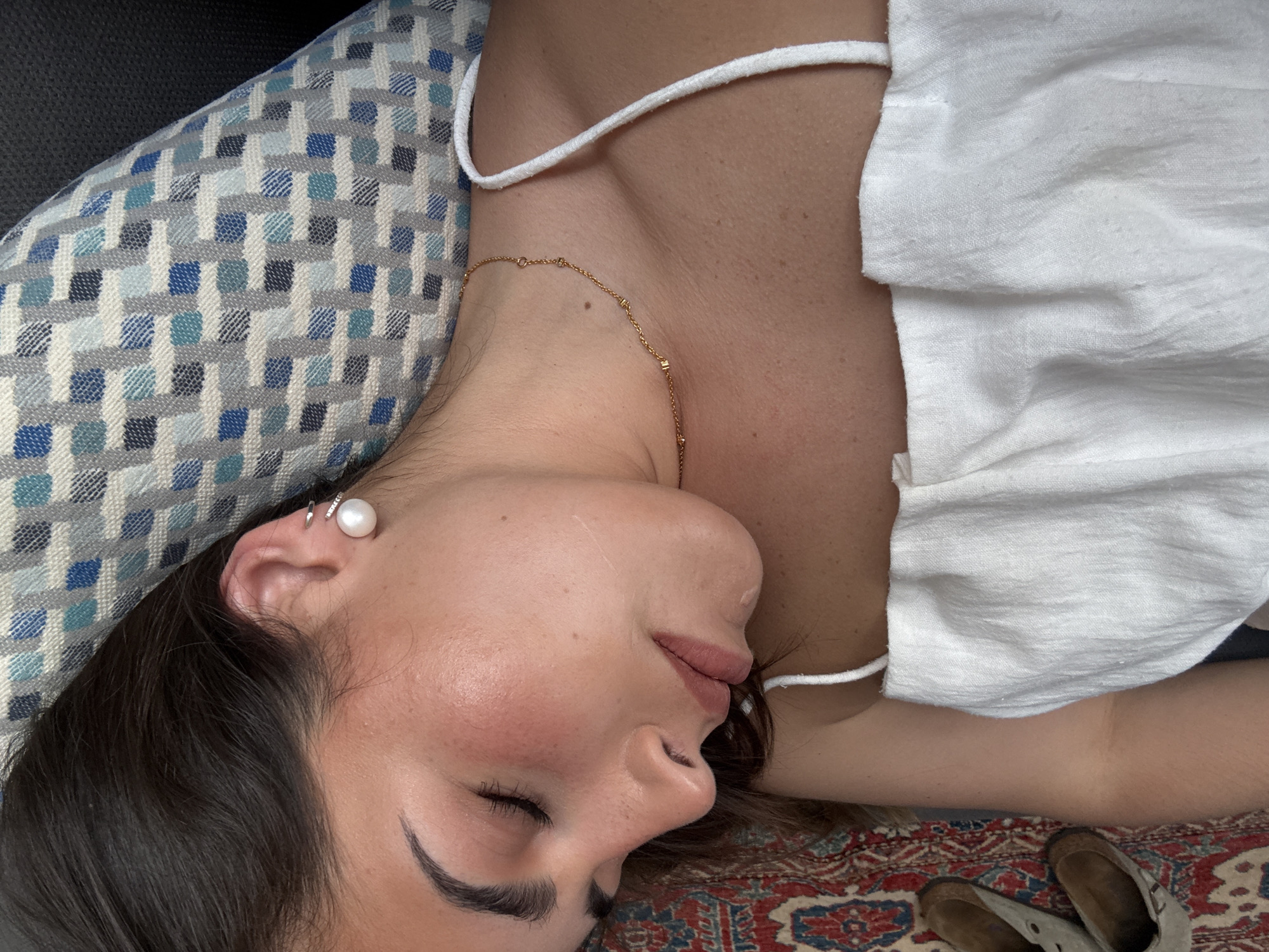 A woman lies with her head on a patterned pillow, wearing a white top and a gold necklace. Her eyes are closed, and she has an earring. Tan shoes are visible on a patterned rug. The location is not specified or discernable from the image.