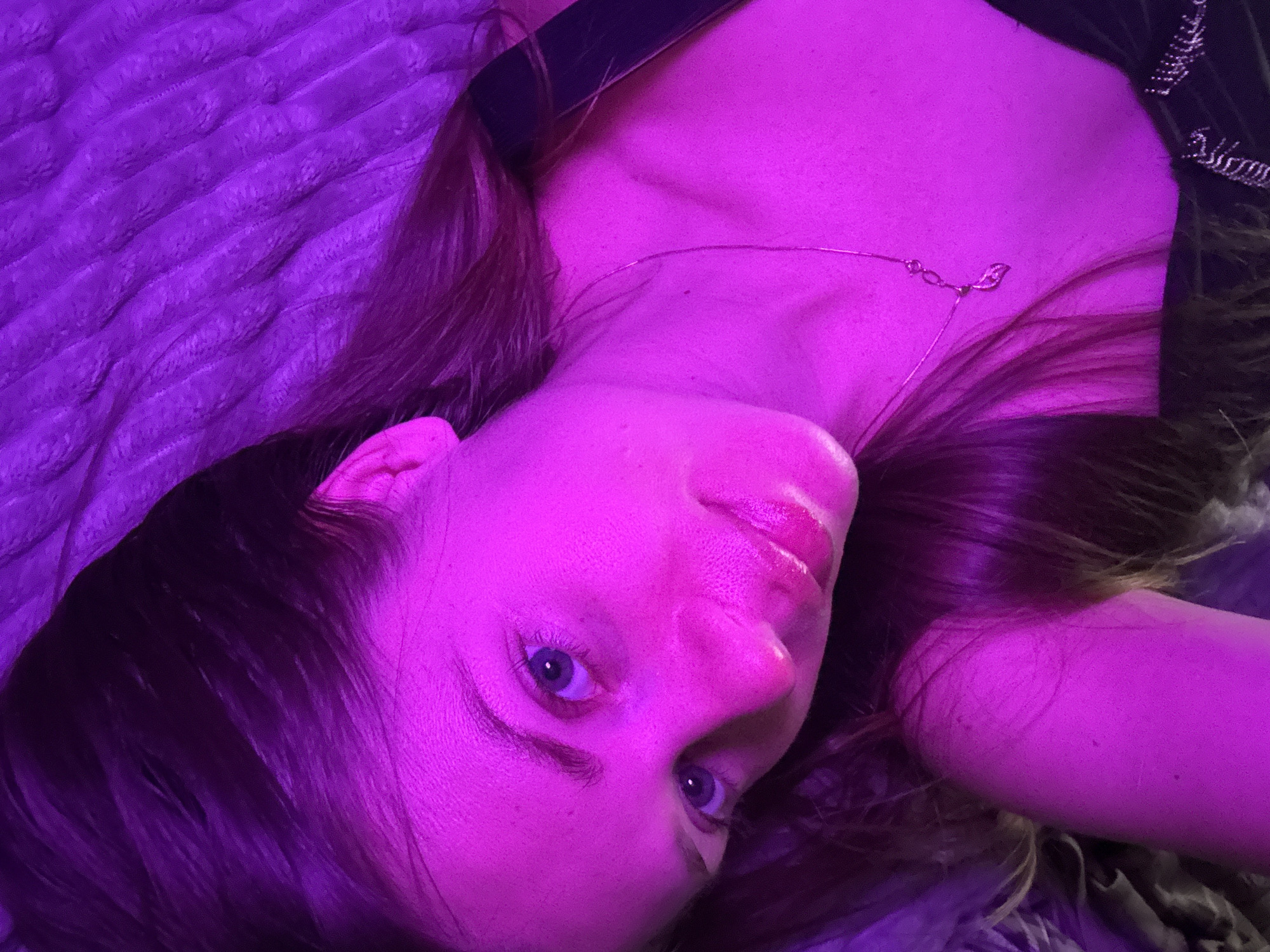 A close-up portrait, taken from a high angle, features a young woman lying down, illuminated by strong magenta-purple lighting that casts a vibrant, uniform hue across the entire scene. Her head is tilted, and she gazes directly at the camera with light-colored eyes, possibly blue. Her full lips appear glossy. She has long, dark hair spread around her head and is wearing a dark top. A delicate necklace with a small, leaf-shaped pendant adorns her neck.
She lies on a textured, quilted surface, likely a bedspread or blanket, which, along with another soft fabric visible on the right, is also bathed in the purple light. The artificial, colored lighting suggests an indoor, nighttime setting. The activity is a posed portrait, with the subject maintaining a calm, direct expression. No text is visible in the image. The provided location context is Batumi, Georgia, though no visual cues in the image confirm this.
