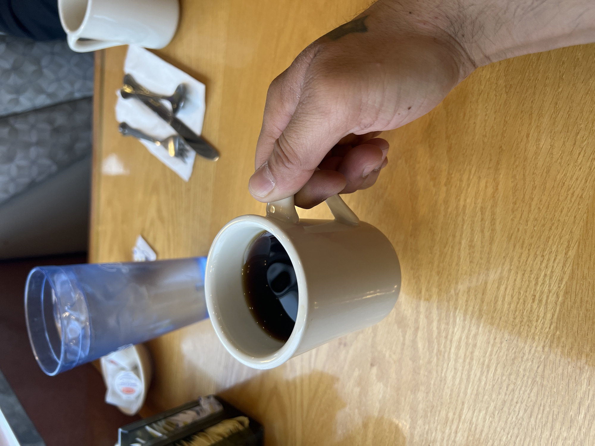 A high-angle shot depicts a hand holding a mug of dark coffee on a wooden table, likely in a restaurant. A water glass and a napkin with silverware are visible on the table. The location is Balmville, United States of America.