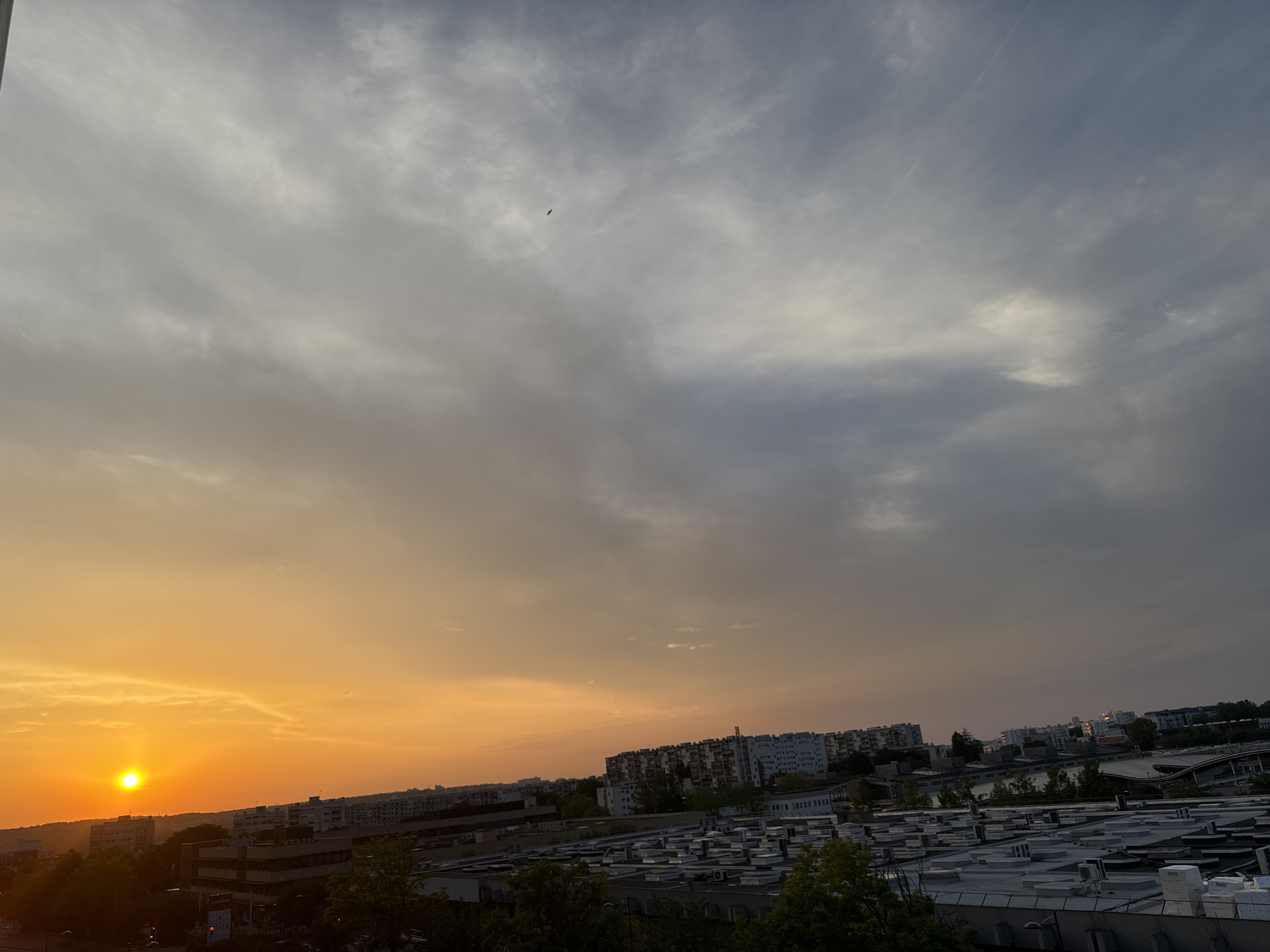 A high-angle shot captures a sunset over a city. The sun, a bright orange orb, is low on the left horizon, casting a warm glow across the sky. The sky transitions from a vibrant orange near the sun to shades of gray and blue as it moves towards the right, with scattered clouds present. Below the sky, the cityscape unfolds, featuring rows of apartment buildings and industrial structures. The buildings vary in height and architectural style, with some appearing to be residential and others possibly commercial or industrial. Many of the buildings have flat roofs, some adorned with air conditioning units or other rooftop equipment. Trees are visible interspersed throughout the urban landscape, with some in the foreground and others further in the background. A small bird is visible in flight in the upper center of the image.