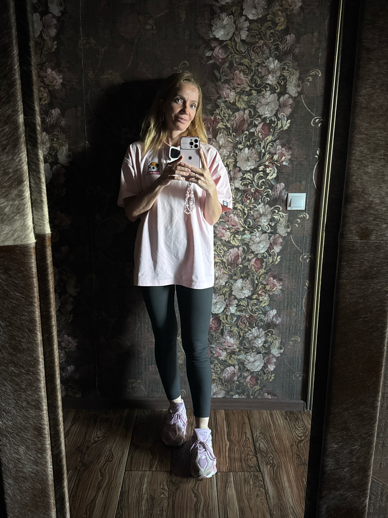 A woman stands in a room, taking a mirror selfie. She is wearing a light pink t-shirt with a cartoon panda graphic, dark leggings, and light purple sneakers. She holds a smartphone in her right hand, angled to capture her reflection. Her left hand is at her side. The background features a dark, floral-patterned wallpaper and a textured brown curtain on the left. The floor is made of wooden planks. A light switch is visible on the wall behind her. The overall lighting suggests an indoor setting, possibly a bedroom or dressing area. There is no indication of the specific city or location within Torzhok, Russia.