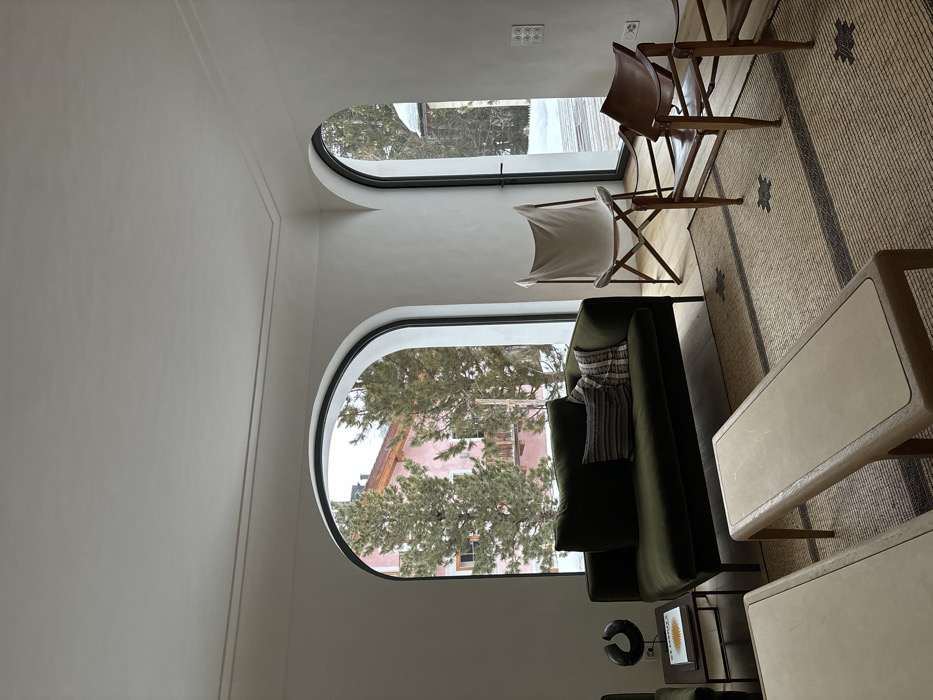 The image shows the interior of a living room in a house located in Celerina/Schlarigna, Switzerland at latitude 46.514, longitude 9.859. The room features a large, green couch placed in front of a large arched window with a view of a snow-covered landscape. The couch is adorned with a throw pillow with a patterned design. To the right of the couch, a wooden armchair with a leather seat and a white canvas chair, folded and placed on the floor, are visible. A brown and white striped rug covers most of the floor in the room. There are two small tables in front of the couch, one of them covered with a book and a black figure. The room has a white ceiling and white walls with a light brown trim. The room is well lit, with natural light streaming in from the arched window. The overall atmosphere is serene and calming.