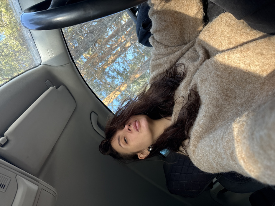The image depicts a female individual seated in the driver's seat of a vehicle, positioned in a natural, wooded environment within the city of Chaikovsky, Russia. The exact location within Chaikovsky is not discernible from the visual evidence. The subject, captured from the chest up, has dark brown, disheveled hair, which falls across her forehead and left shoulder. She wears a light brown, oversized knit sweater and a silver-colored hoop earring in her left ear. Her head is slightly tilted, and her gaze is directed towards the camera with a neutral expression. The vehicle's interior features a light-colored headliner, a folded sun visor on the passenger side, and a portion of a black steering wheel to the right. An "AIRBAG" label is visible on the passenger side A-pillar. The windshield is covered with numerous small water droplets or condensation. Outside the vehicle, a clear blue sky is visible above a sparse canopy of bare trees, indicating a cold season. The scene is illuminated by strong, natural sunlight.