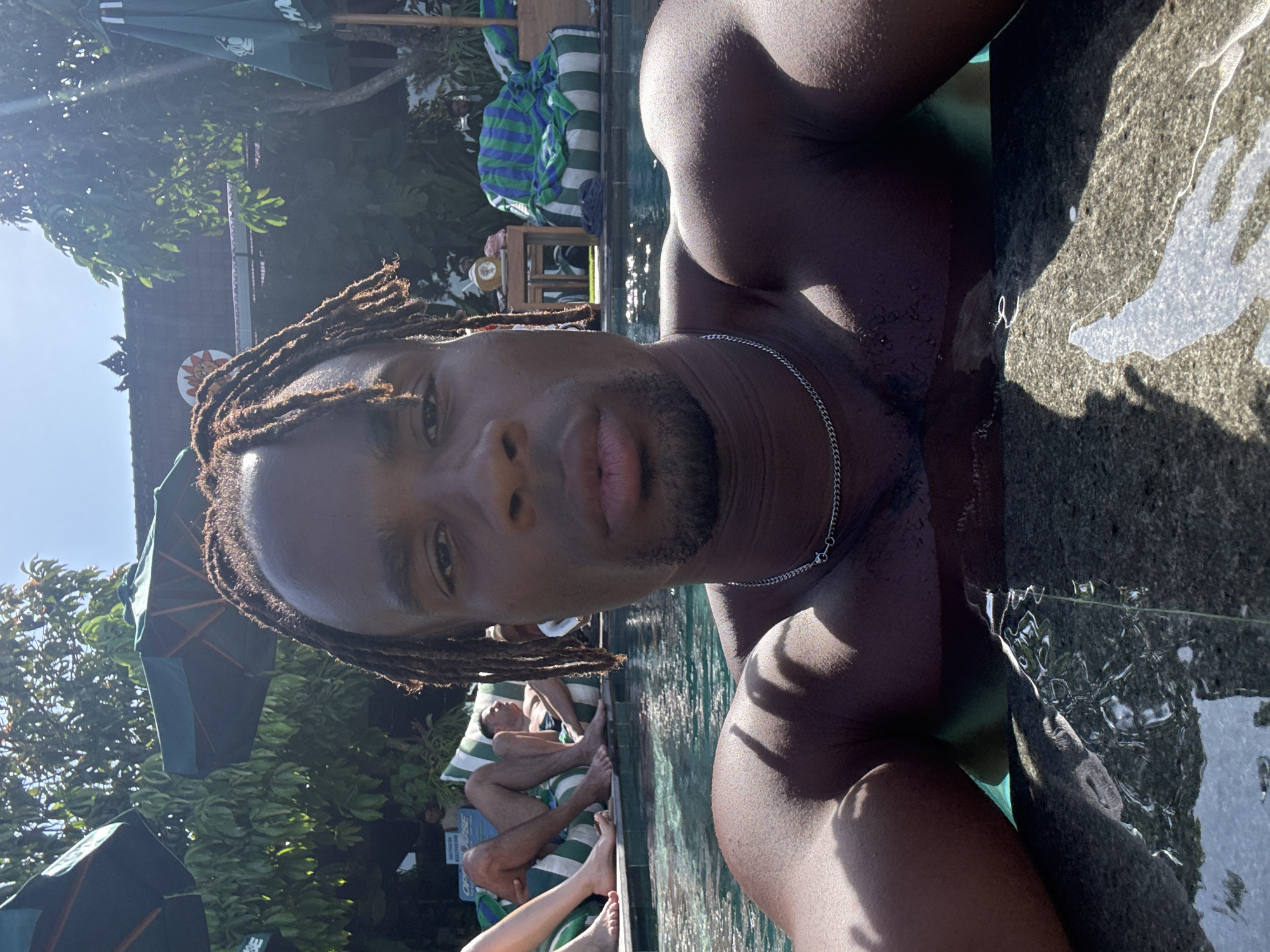 A close-up shot captures a man with dreadlocks and a silver chain standing in a pool. The pool appears to be located in a tropical resort, judging by the surrounding greenery and sun umbrellas. Other people are lounging on chairs in the background. The specific location is unknown.