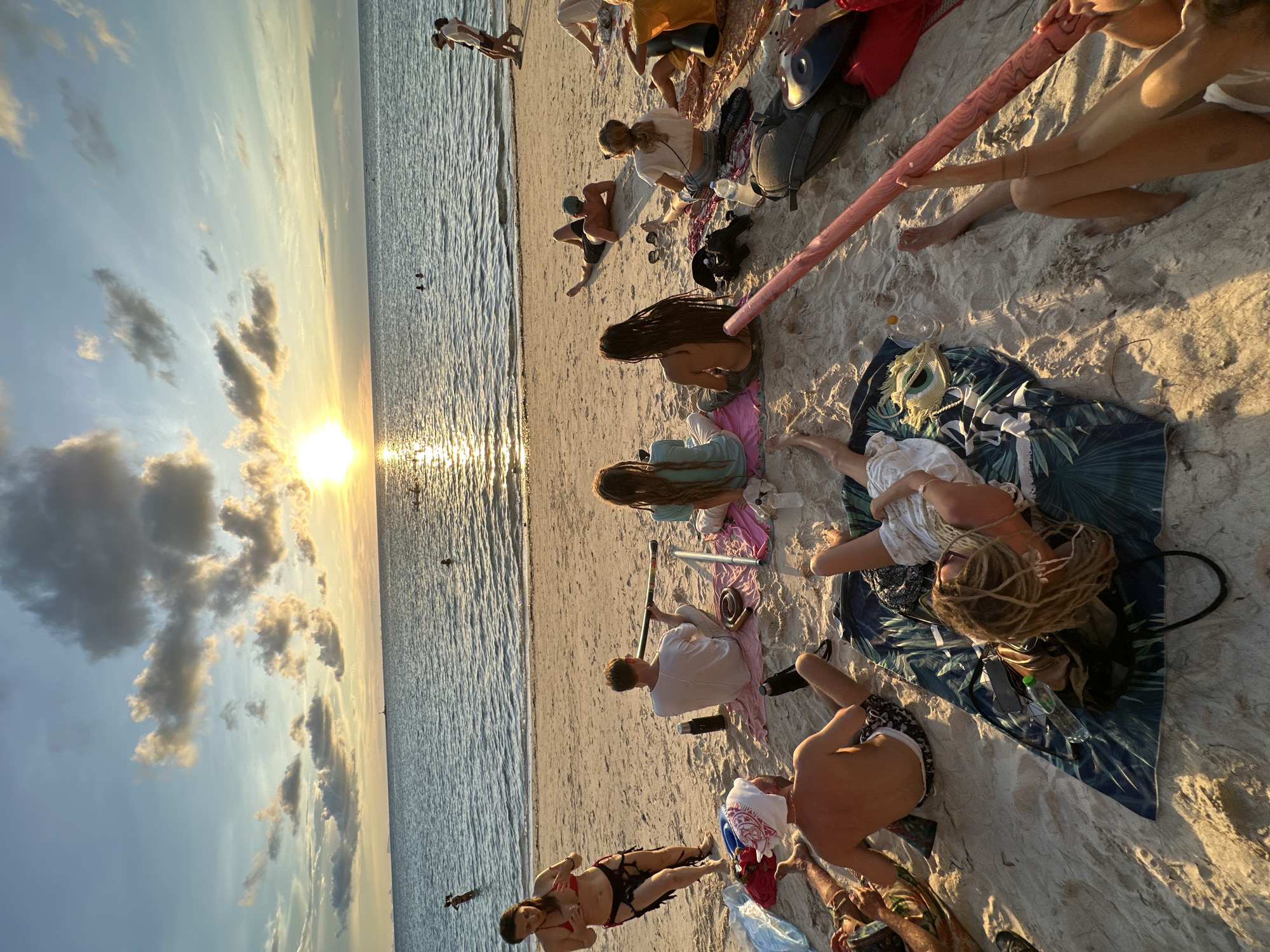 At sunset on a beach in Koh Phangan, Thailand, a group of people are gathered on the sand. The sky is a mix of blue and clouds, with the sun low on the horizon, casting a golden light on the water and sand. The sea is calm, with gentle waves lapping at the shore.
Several individuals are seated on the sand, some on blankets. One person is playing a didgeridoo. Another person, a woman in a red bikini, stands near the water's edge. A few other people are visible in the water, swimming or wading.
Objects scattered on the sand include towels, water bottles, and what appears to be a hang drum. The overall atmosphere is relaxed and communal, with the setting sun providing a beautiful backdrop.