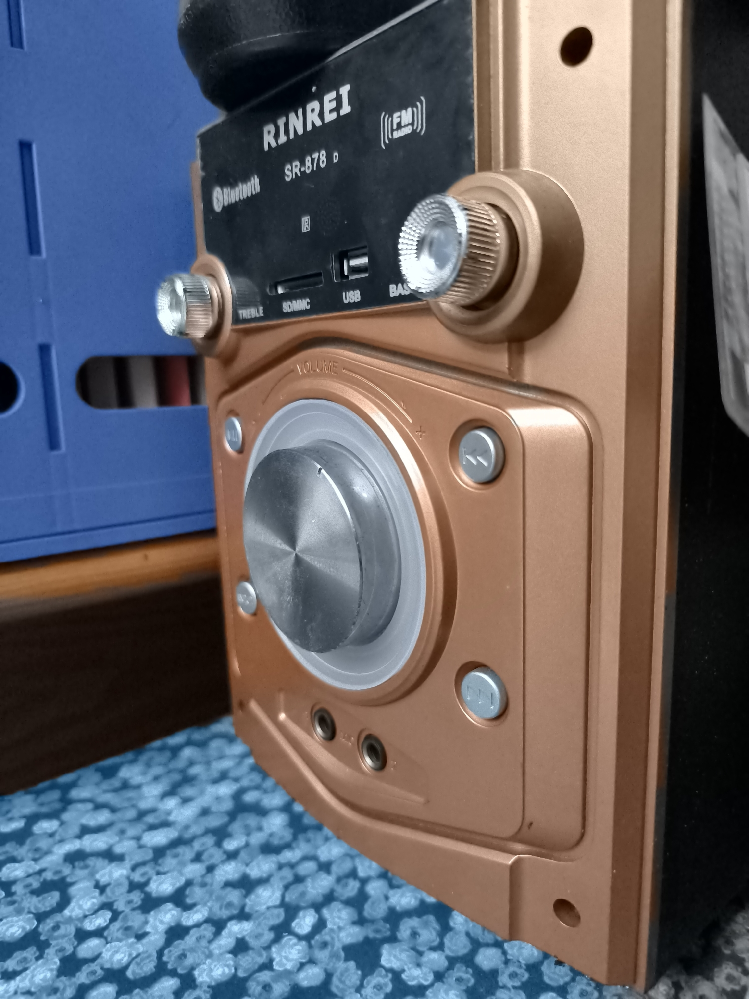 A close-up, eye-level, slightly angled shot shows the front panel of a portable speaker unit. The speaker is made of a copper-colored plastic with metallic silver accents. The top of the speaker has a black, textured surface. The front panel features various controls and inputs. A large silver dial labeled "VOLUME" is prominent in the center. To the left and right of this dial are smaller silver knobs, one labeled "TREBLE" and the other "BASS." Below the "TREBLE" knob are inputs for "SD/MMC" and "USB." Above these, the brand name "RINREI" is printed in white, followed by "SR-878 D". To the right of the brand name, an "FM RADIO" icon is displayed. Several small silver buttons are also visible on the panel. The speaker unit is positioned next to a blue plastic container, and a blue patterned cloth is visible in the foreground. The overall impression is of a consumer electronics device on a patterned surface. The scene is located indoors.